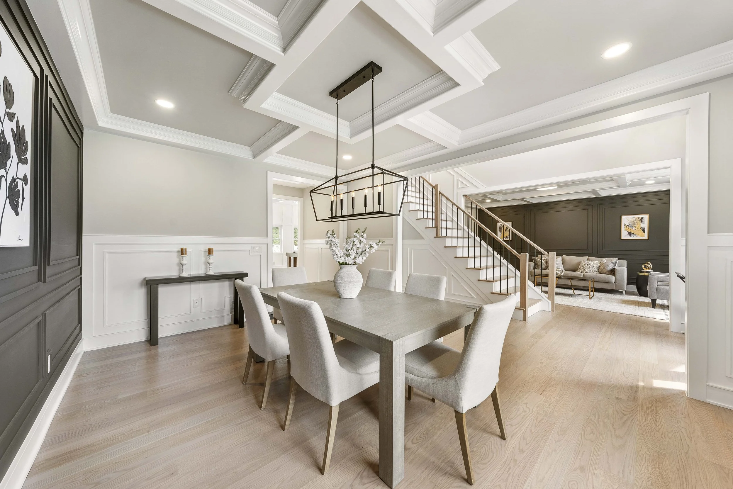 Modern dining room with light wood flooring, a wooden table, and eight beige upholstered chairs. A black geometric chandelier hangs above the table, and a white vase with white flowers is on the table. There is a black wainscot with white walls and i