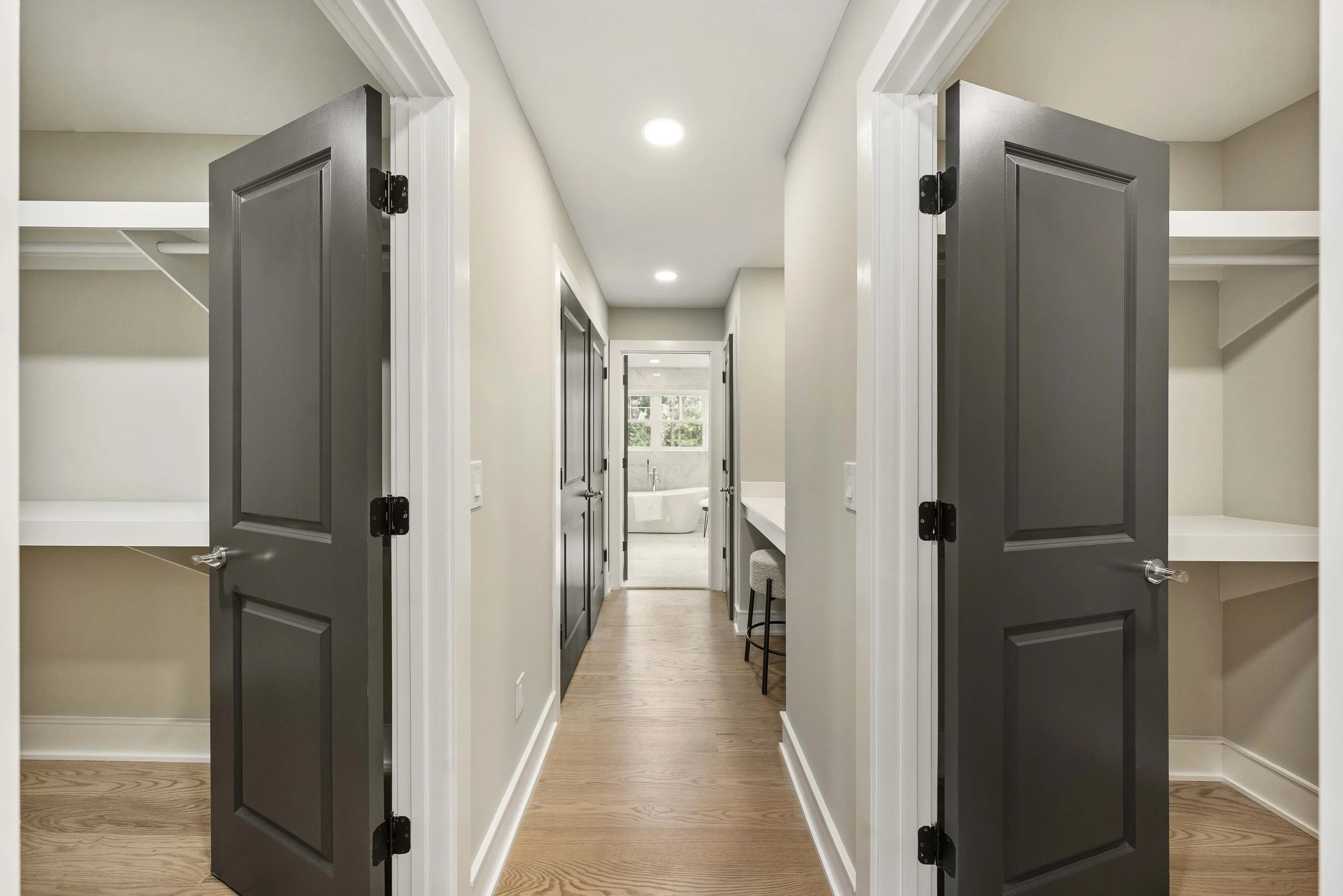 A hallway with two open closets on each side, dark gray doors, light-colored walls, hardwood flooring, leading to a bathroom with a bathtub at the end and natural light coming through windows.