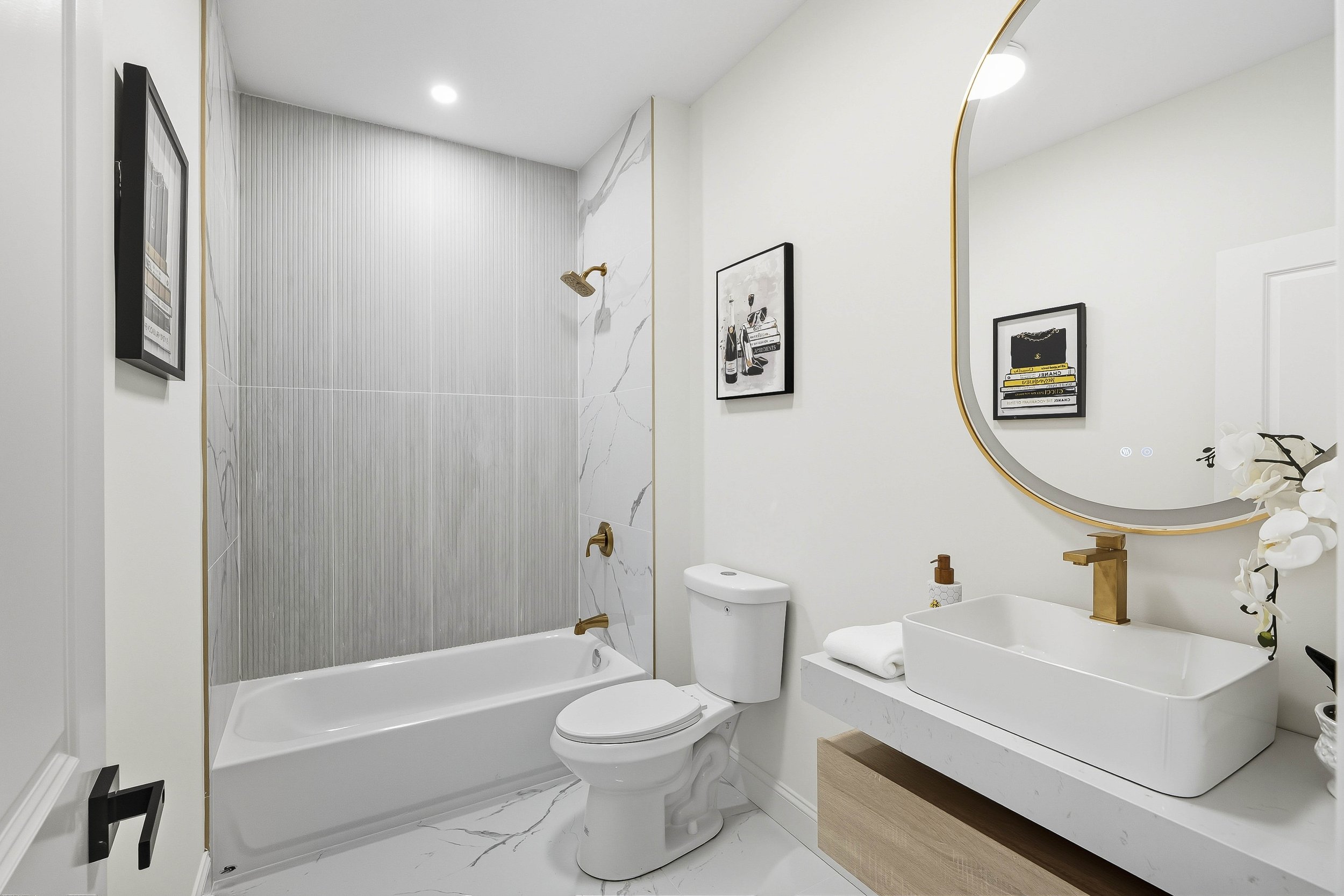 Modern bathroom with white walls, marble flooring, a bathtub with gold fixtures, a toilet, a vanity with a rectangular sink, black and white artwork, a large mirror with a gold frame, and decorative items.