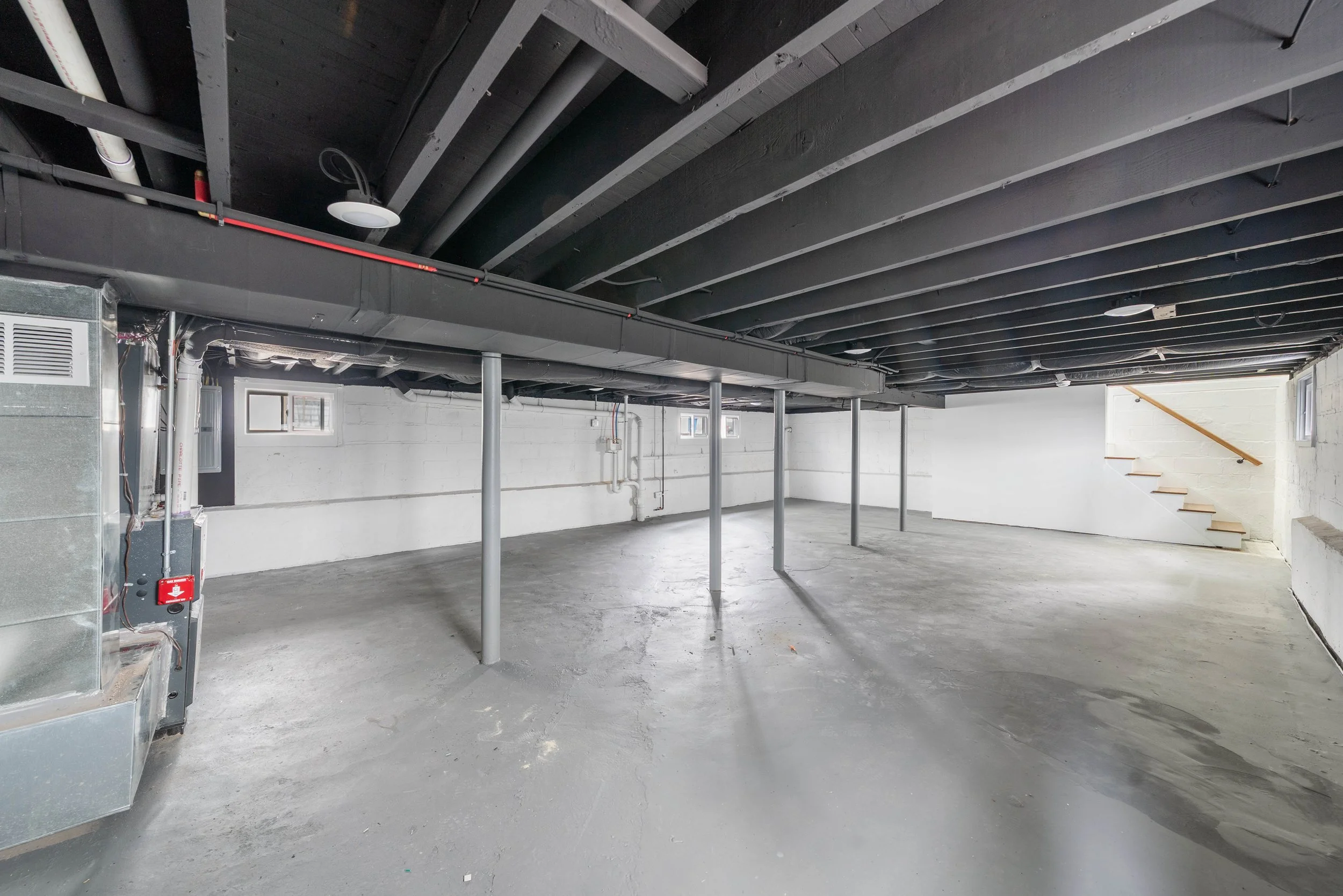 Empty unfinished basement with concrete floor, white walls, black ceiling with exposed beams and ducts, small windows, and a staircase at the far end.