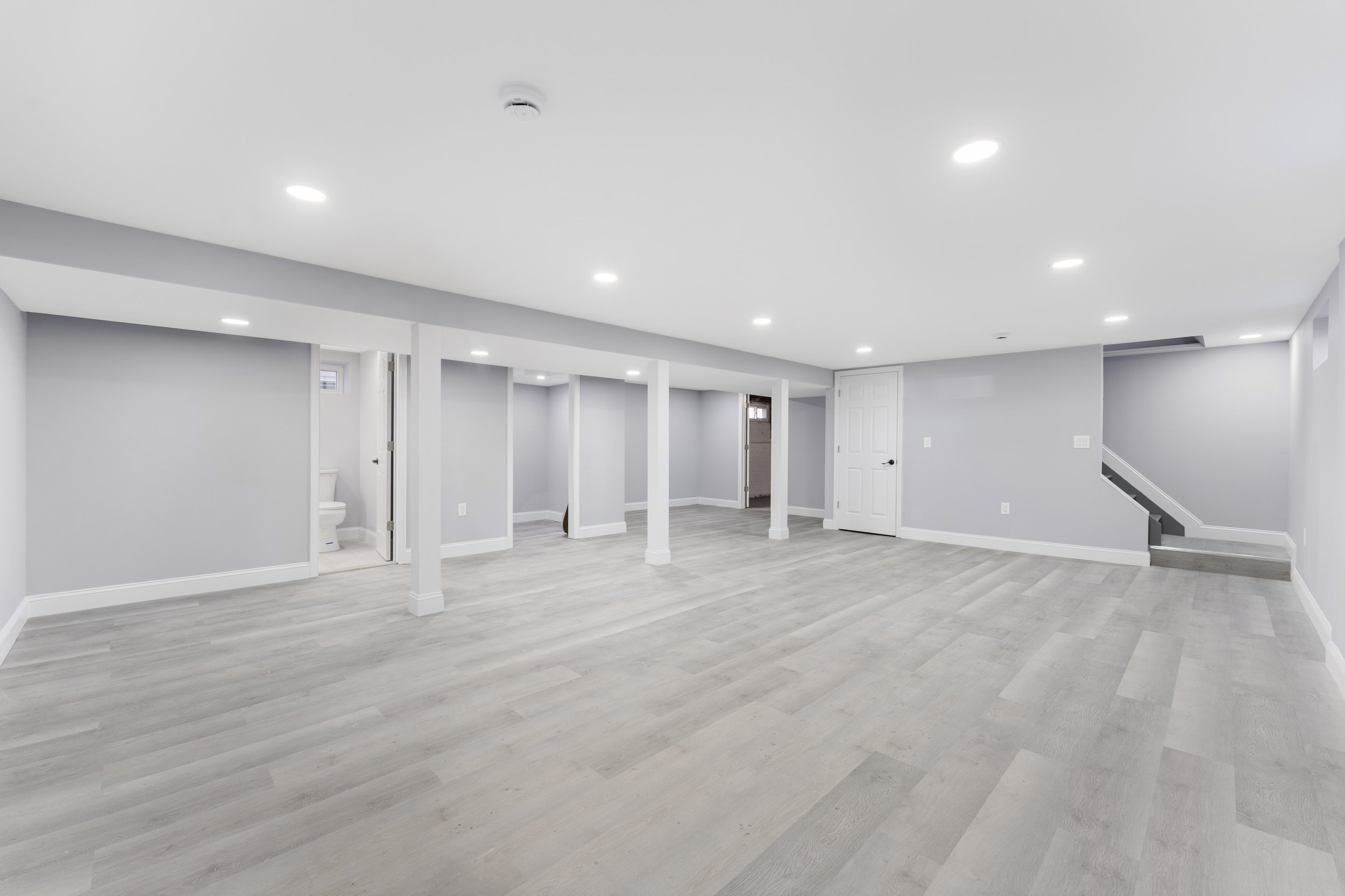 Empty finished basement with light gray walls, white ceiling, wood-look gray flooring, and six recessed lights, including a small bathroom in the back left corner.