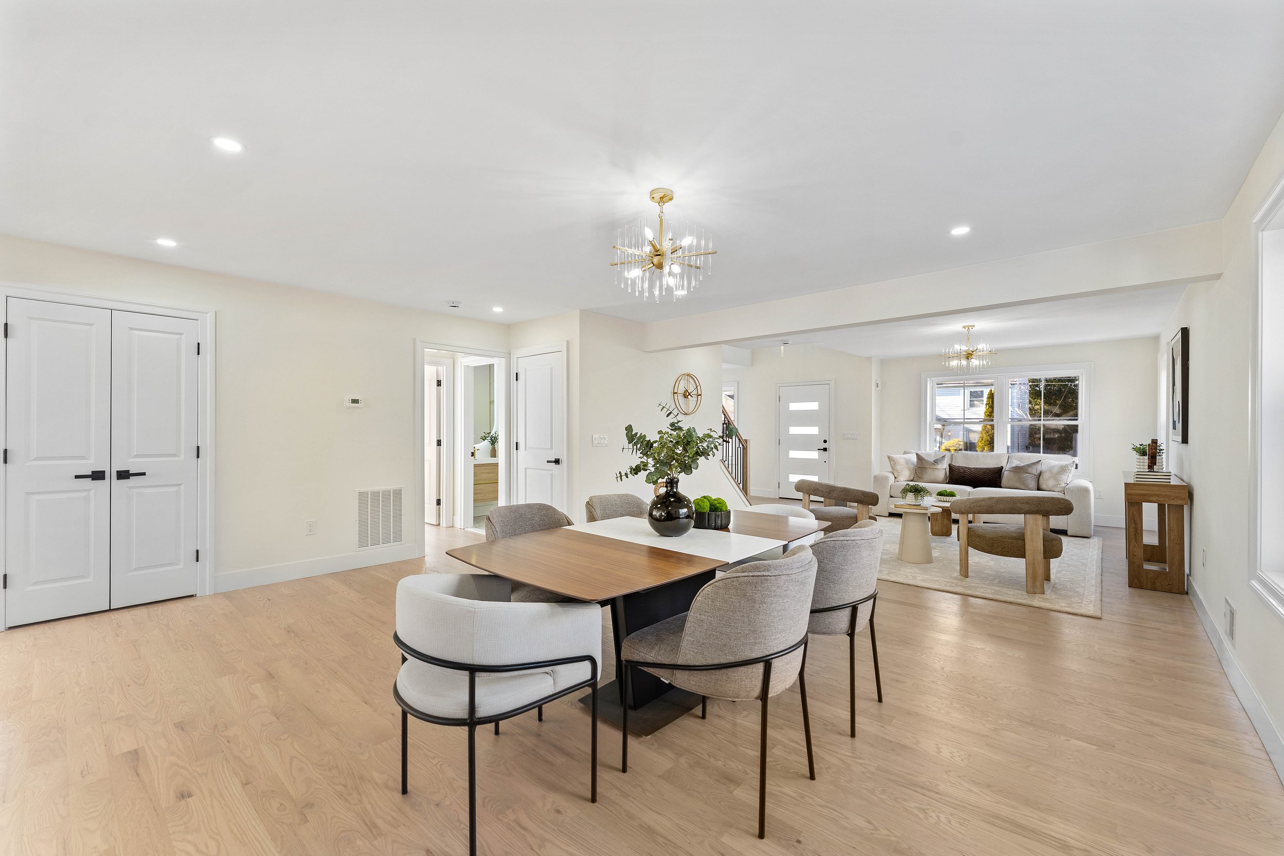 Open-concept living and dining room with hardwood floors, white walls, modern chandelier, dining table with six upholstered chairs, decorative vase with greenery, and a cozy sitting area with a sofa and armchairs near large windows.