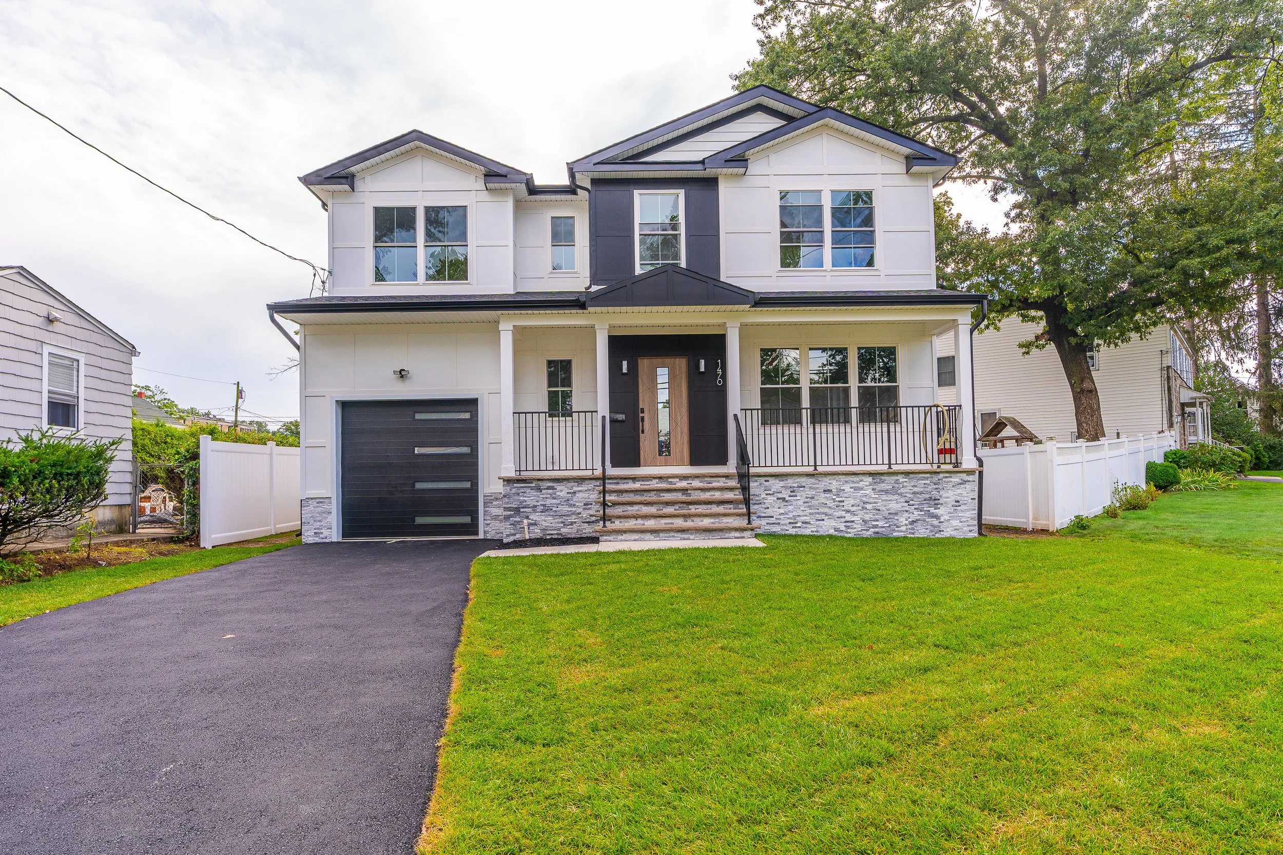 Front view of a modern two-story house with black and white exterior, black garage door, front porch with stairs, and well-maintained lawn with trees and white fence.