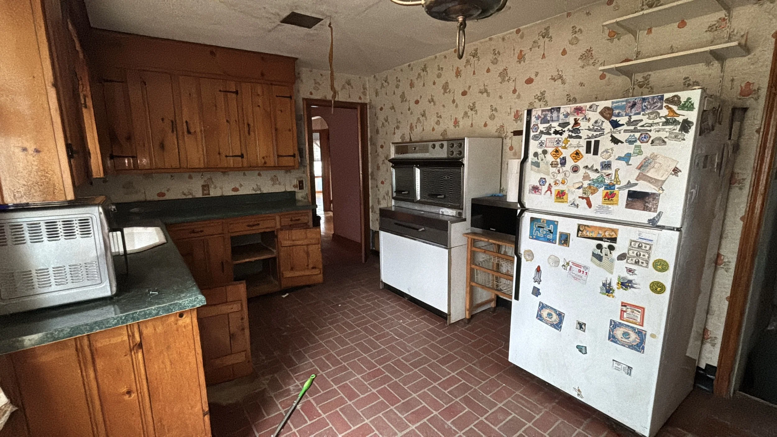 A vintage kitchen with wooden cabinets, a dark green countertop, a white and black refrigerator covered in stickers, a metal microwave, and a brick floor. The wallpaper features small orange and green floral patterns.