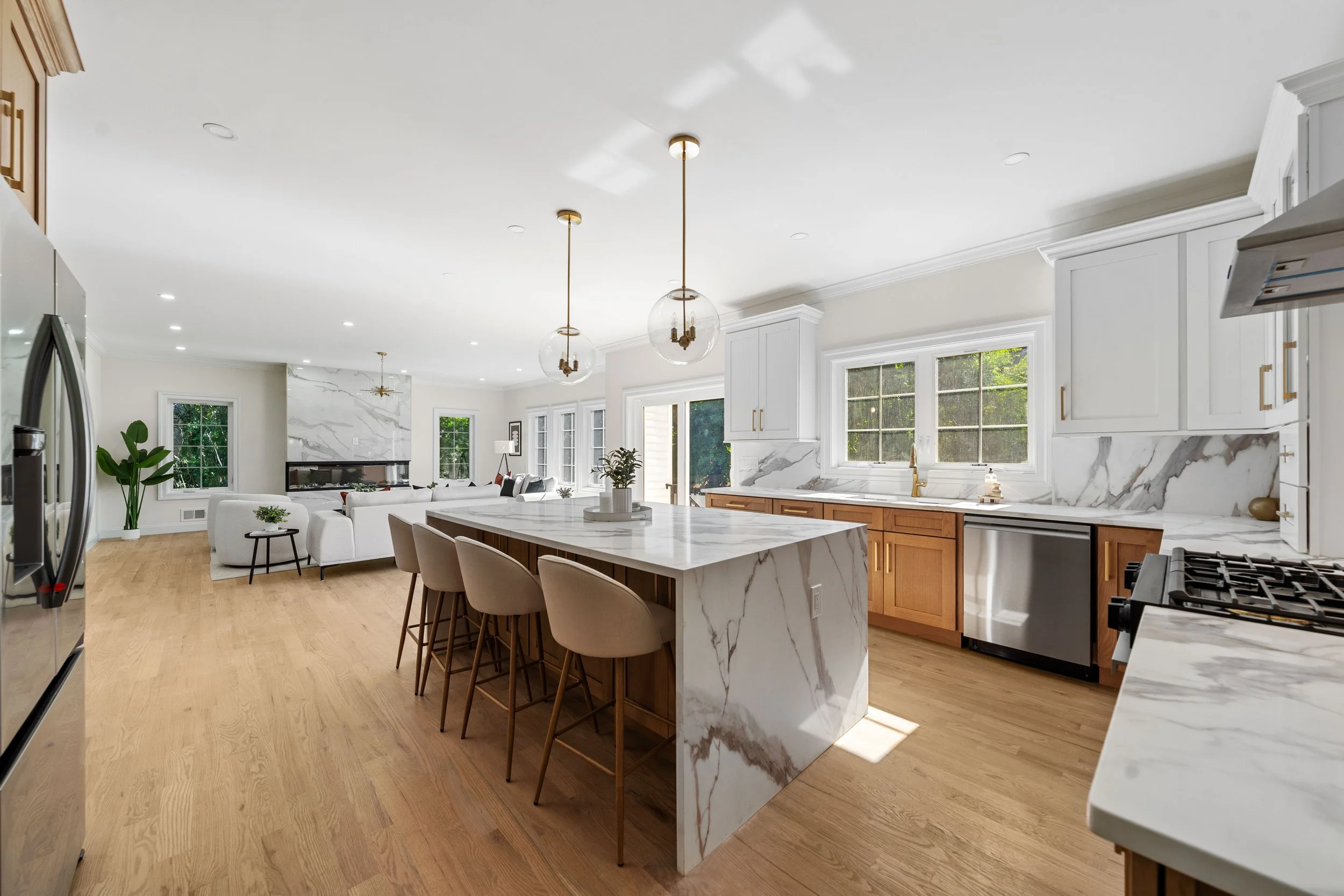 Modern open-concept kitchen with white cabinetry, marble countertops, a large island with beige barstools, stainless steel appliances, and hardwood floors, opening into a living room with white furniture and large windows.