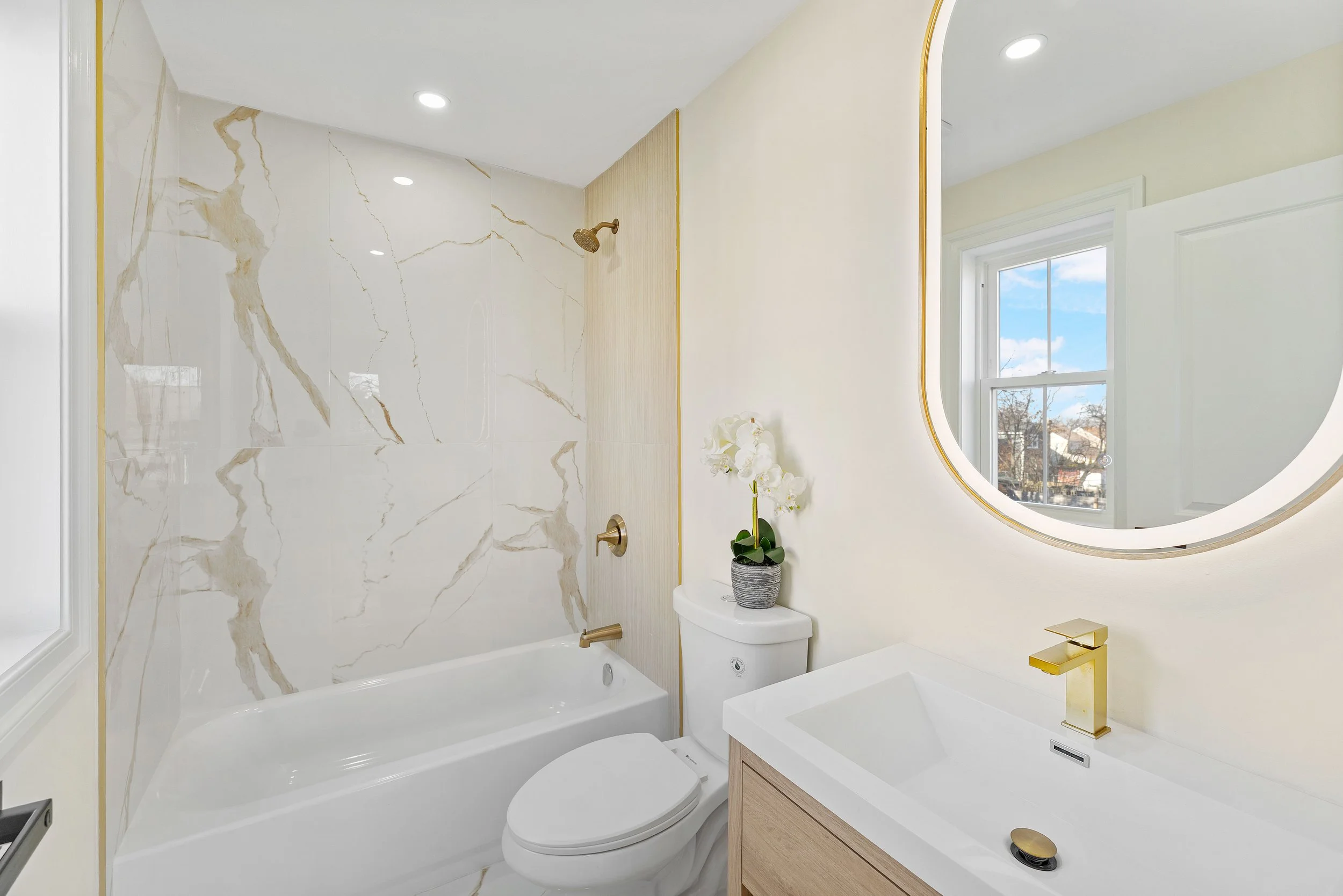 Modern bathroom with marble shower wall, white bathtub, toilet, white sink with gold faucet, large mirror, window showing a blue sky and trees outside, and a potted white orchid on top of the toilet tank.