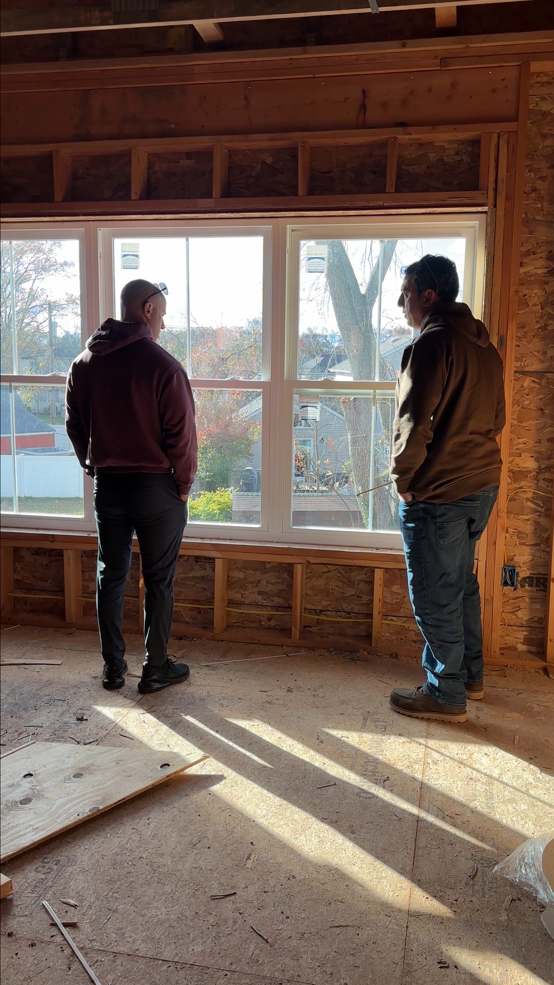 Two men standing inside a house under construction, looking out a large window, with sunlight streaming in.