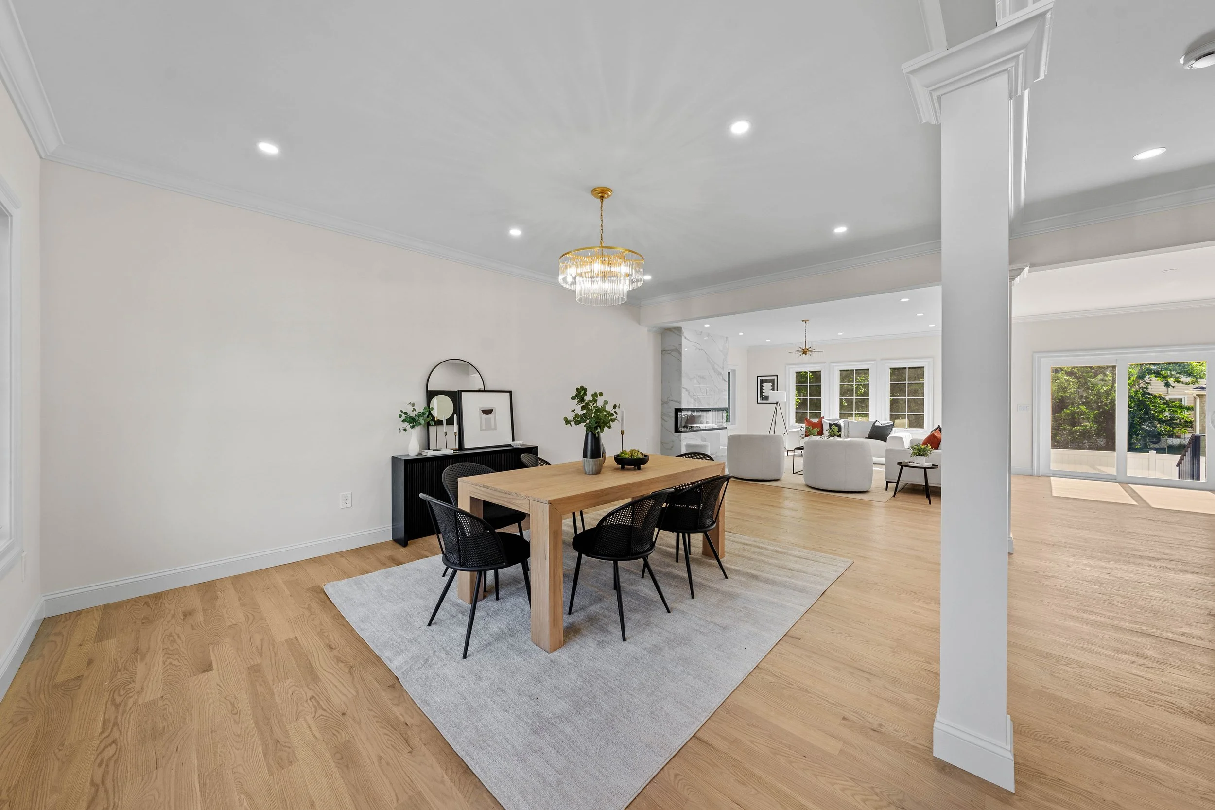 Open-concept living and dining area with light hardwood floors, white walls, modern chandelier, and ample natural light from large windows.