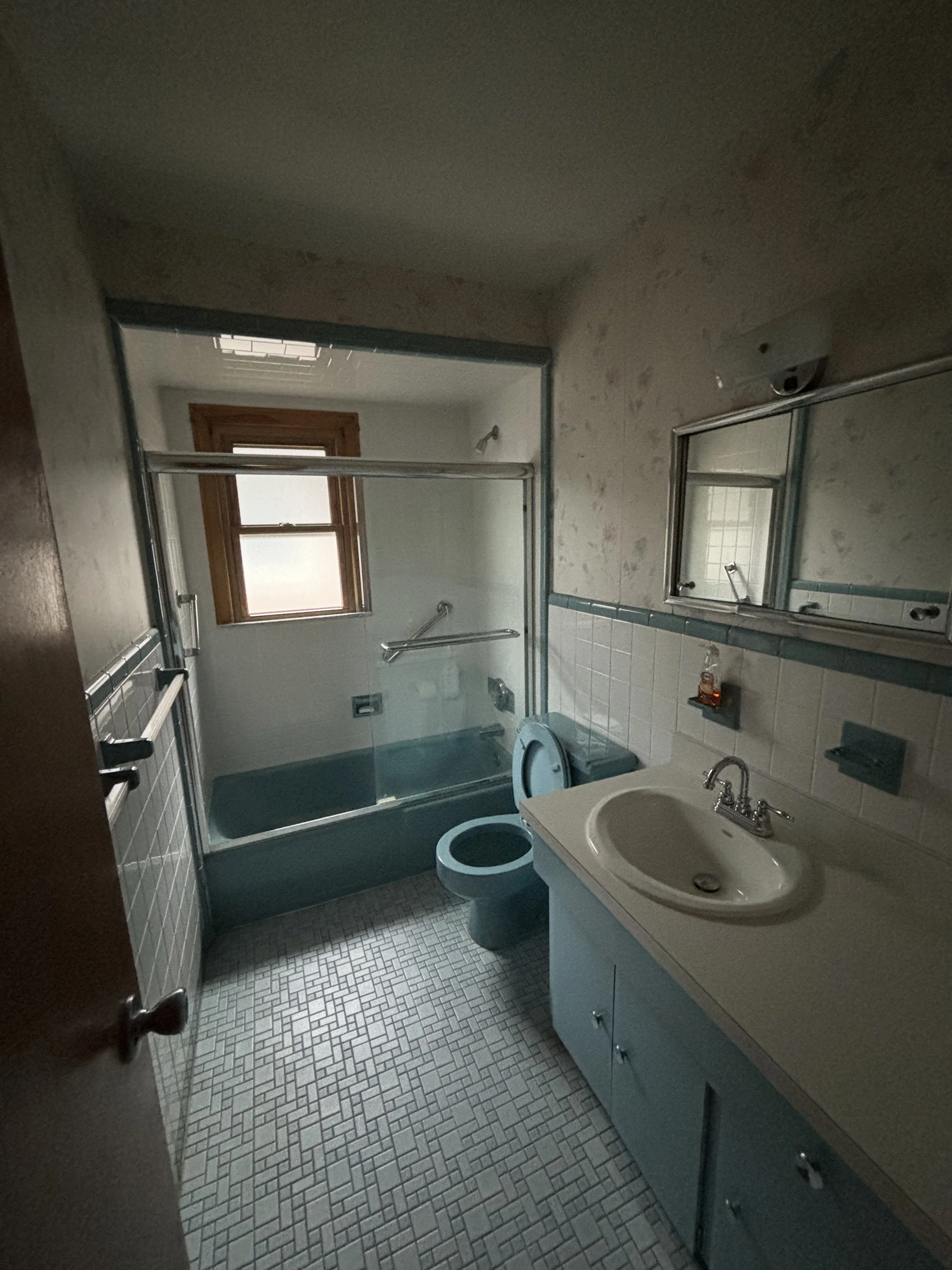 A small bathroom with a shower, toilet, and sink. The shower has a glass door, and there is a window above it. The walls are partially tiled, and the floor has small square tiles.