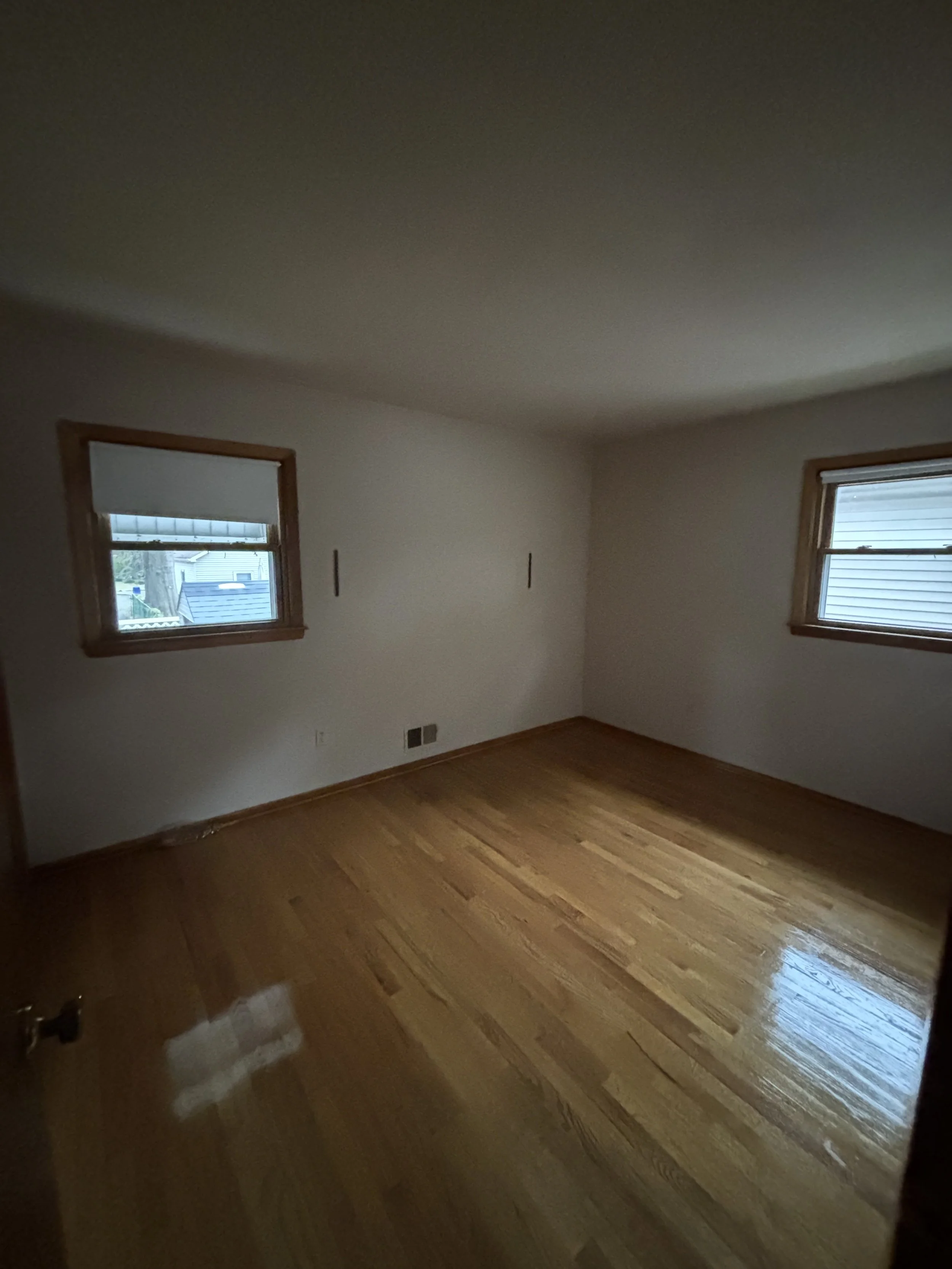 Empty room with hardwood floors, two windows with blinds, plain white walls, and wooden trim.