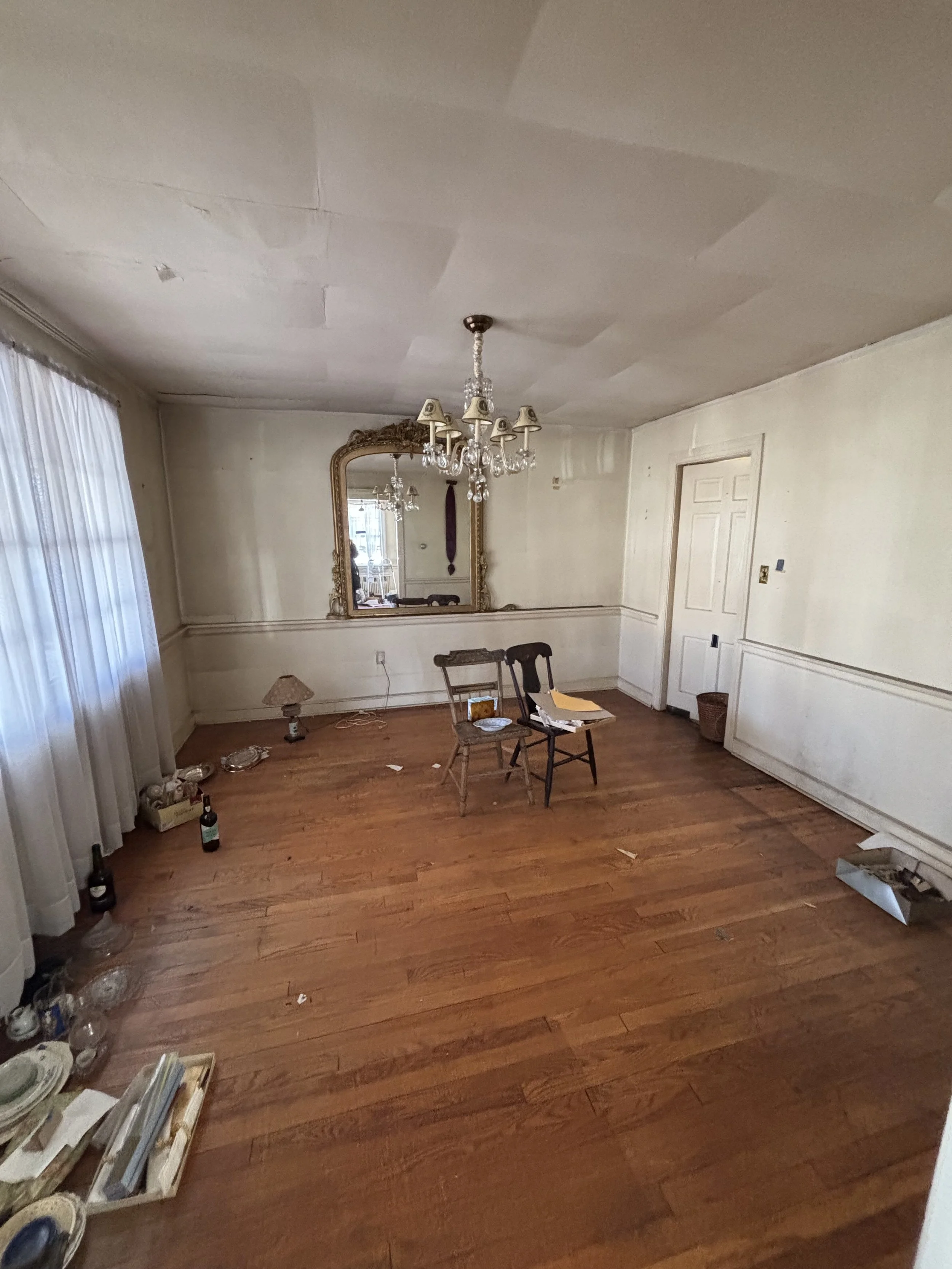 Empty living room with hardwood floor, chandelier, mirror, and scattered items including bottles, dishes, and papers.