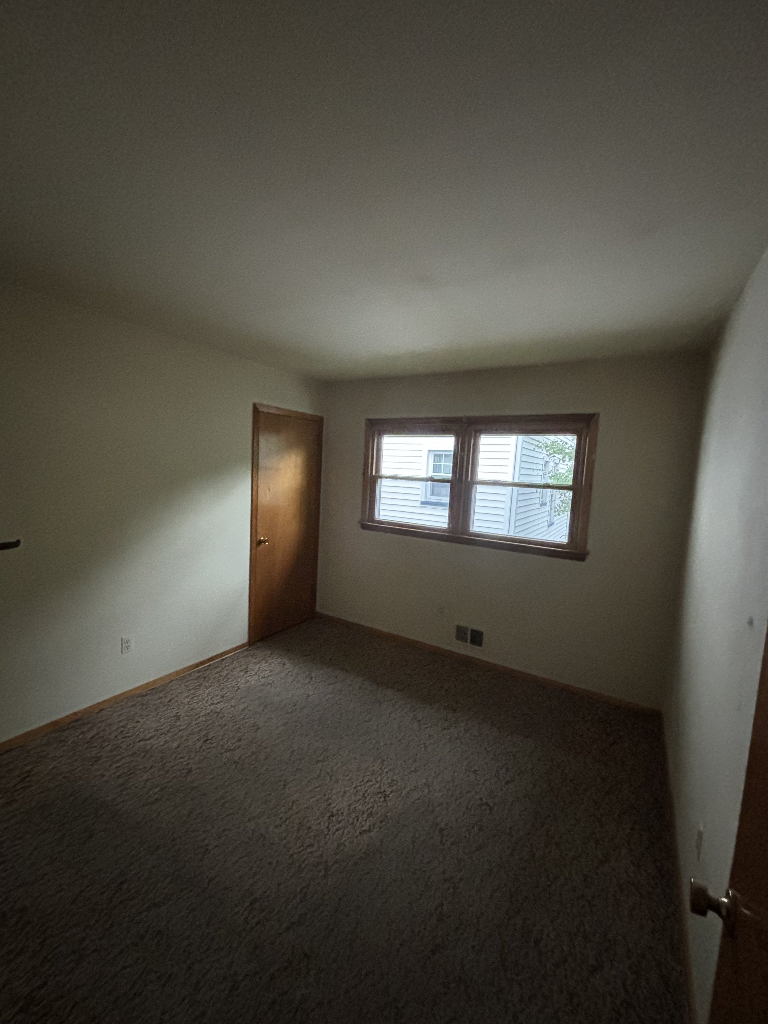 Empty bedroom with beige carpet, white walls, a wooden door, and two windows showing neighboring houses outside.