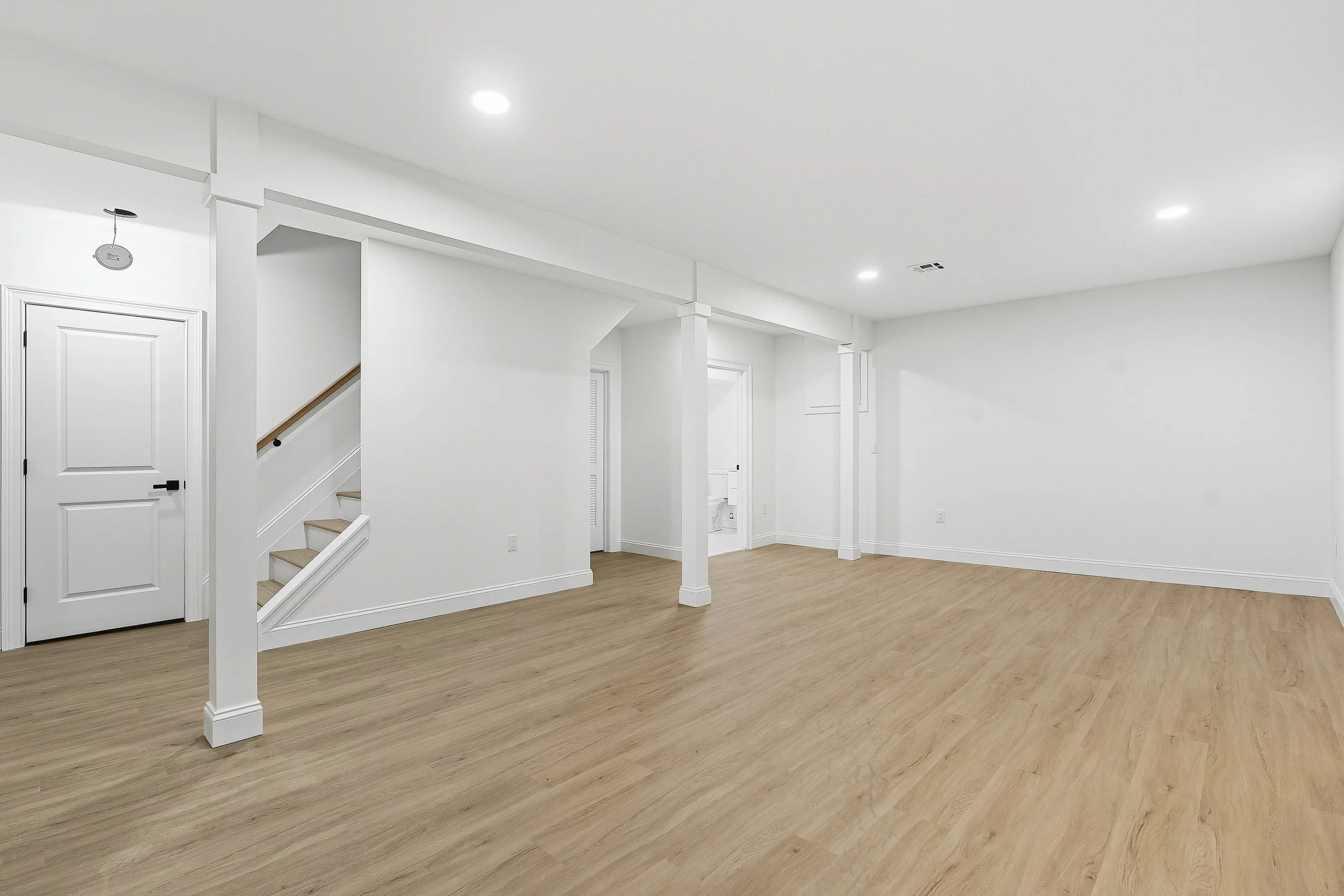 Empty room with white walls, wooden flooring, a staircase with wooden steps and a white railing, and a closed white door. Recessed ceiling lights illuminate the space.