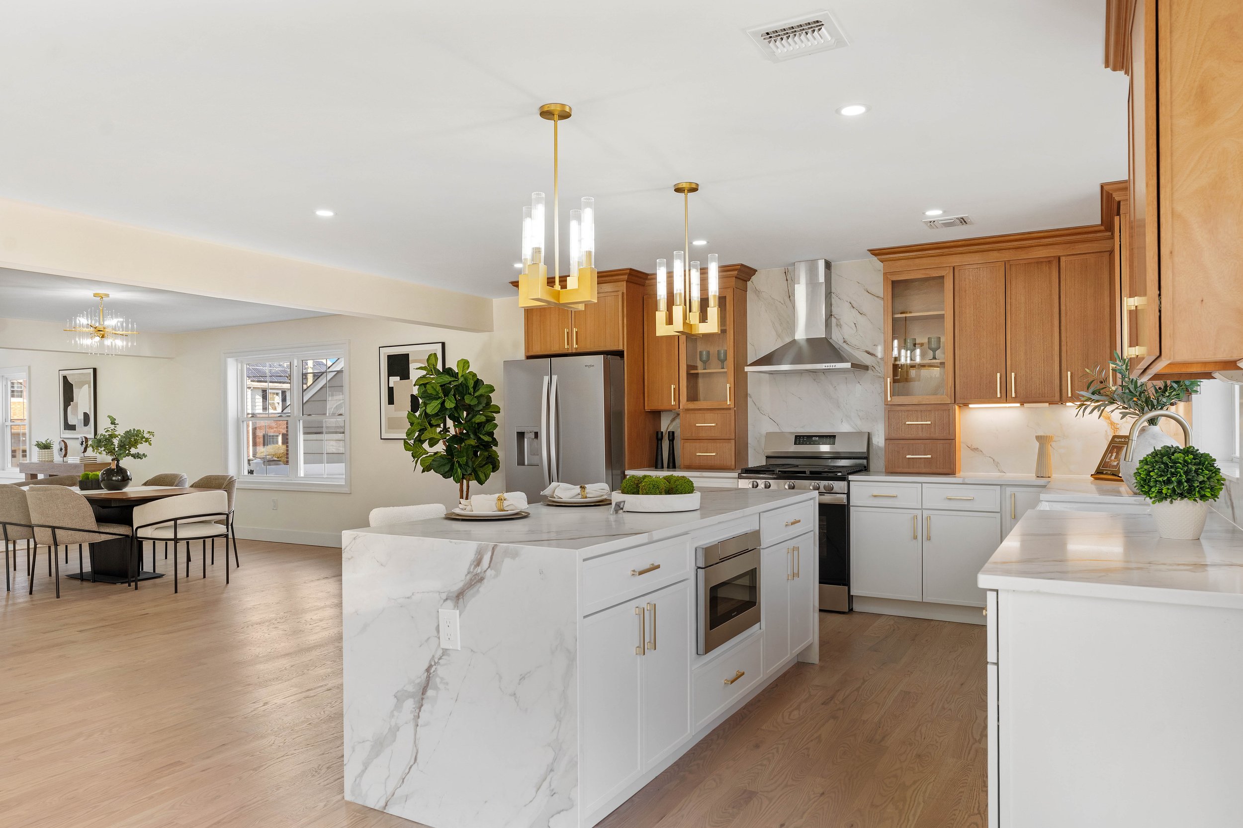 Modern kitchen with white marble countertops, wooden cabinets, stainless steel appliances, and a kitchen island with green plants, in an open-concept living space with dining area.