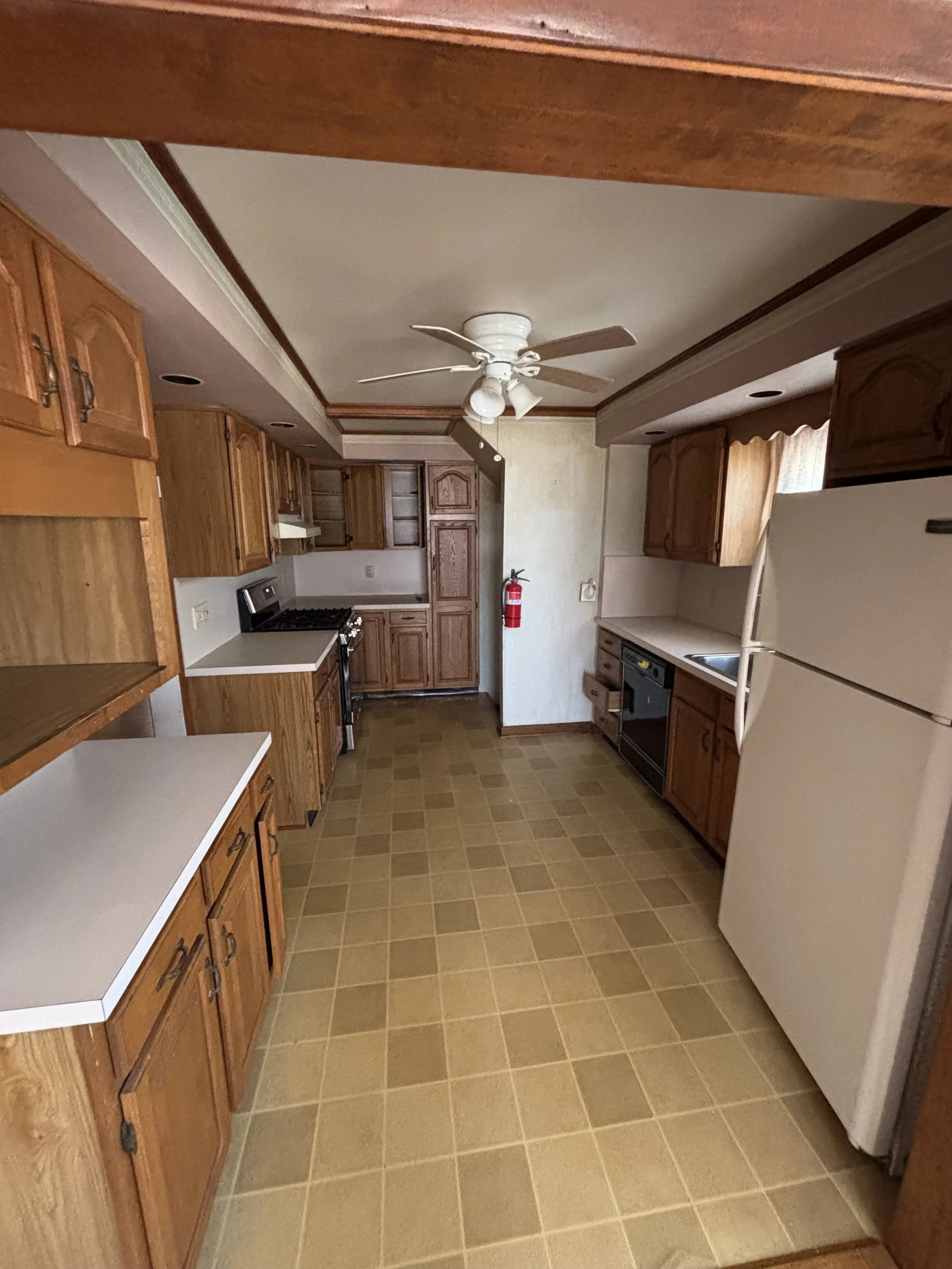 A kitchen with wooden cabinets, white countertops, a white refrigerator, a black stove, and a ceiling fan. Fire extinguisher is mounted on the wall, with a window above the sink.