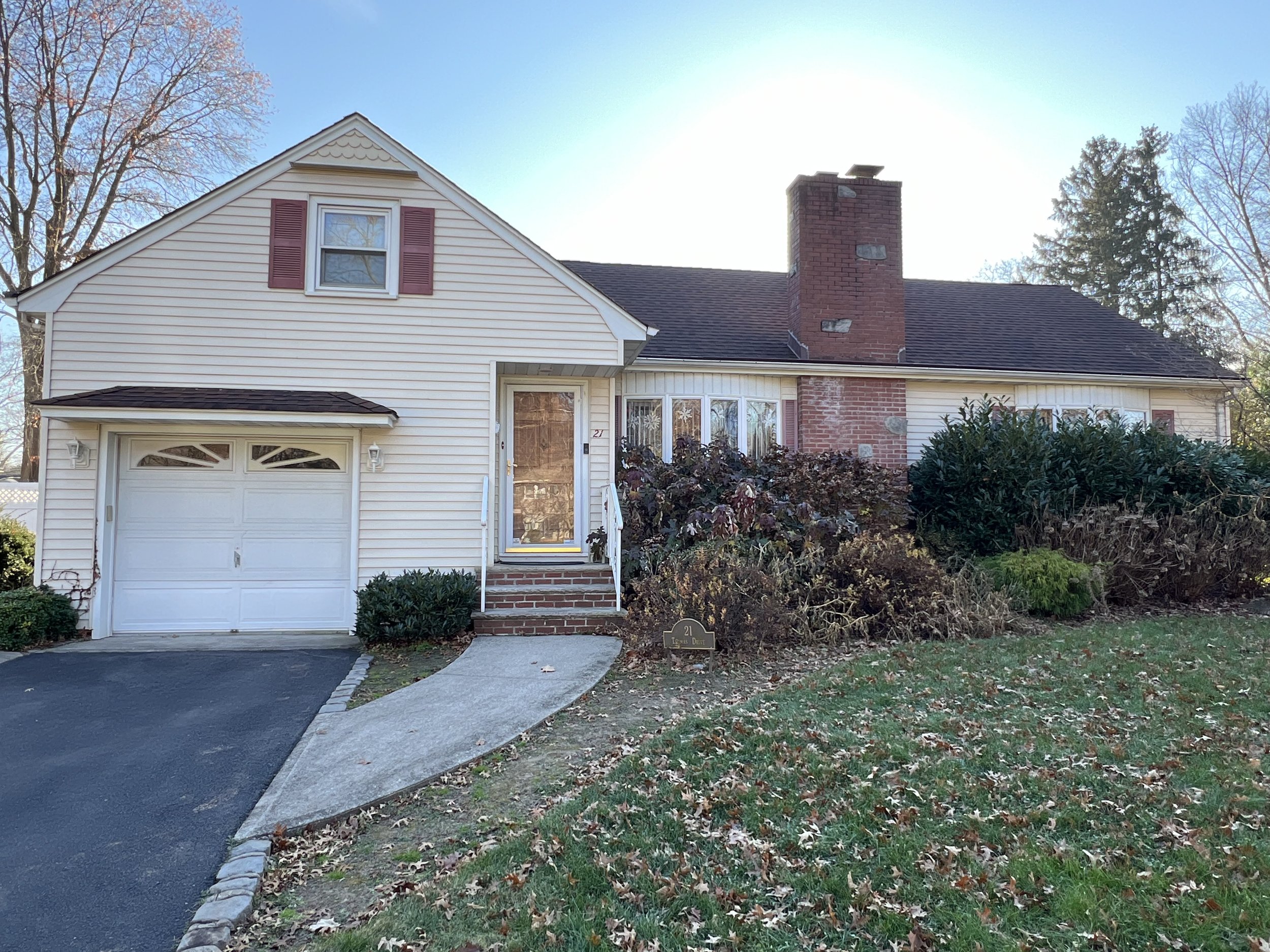 Front view of a suburban house with a white garage door, brick chimney, and a small front porch with a glass door. The house has cream-colored siding with red shutters, and the front yard has a lawn with fallen autumn leaves and trimmed bushes.