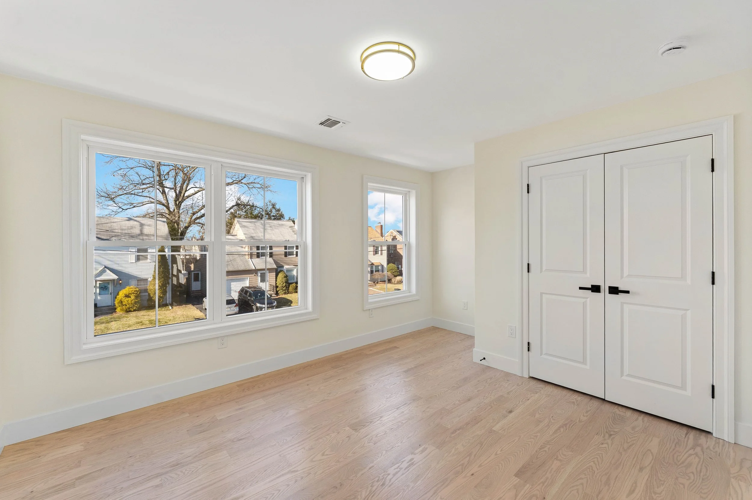 Empty bedroom with light-colored walls, hardwood floors, large windows, white closet doors, and ceiling light fixture.