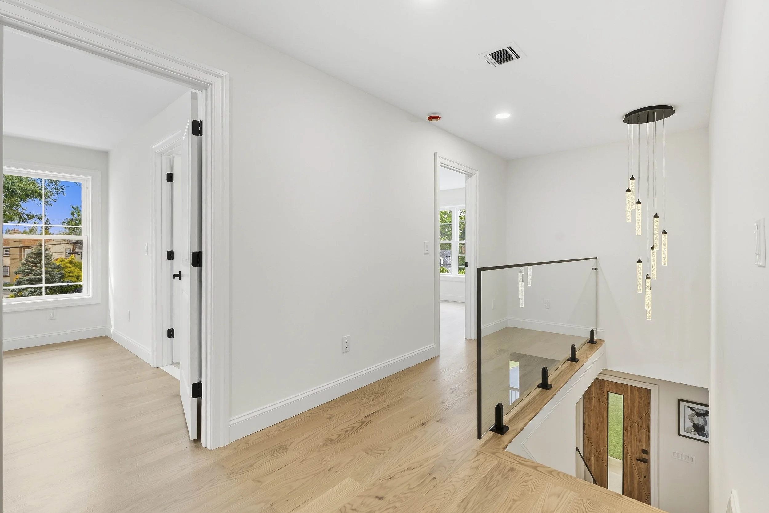 Interior view of a modern house with white walls, hardwood floors, and a staircase with glass and black railing, pendant lighting, and multiple rooms visible through doorways.