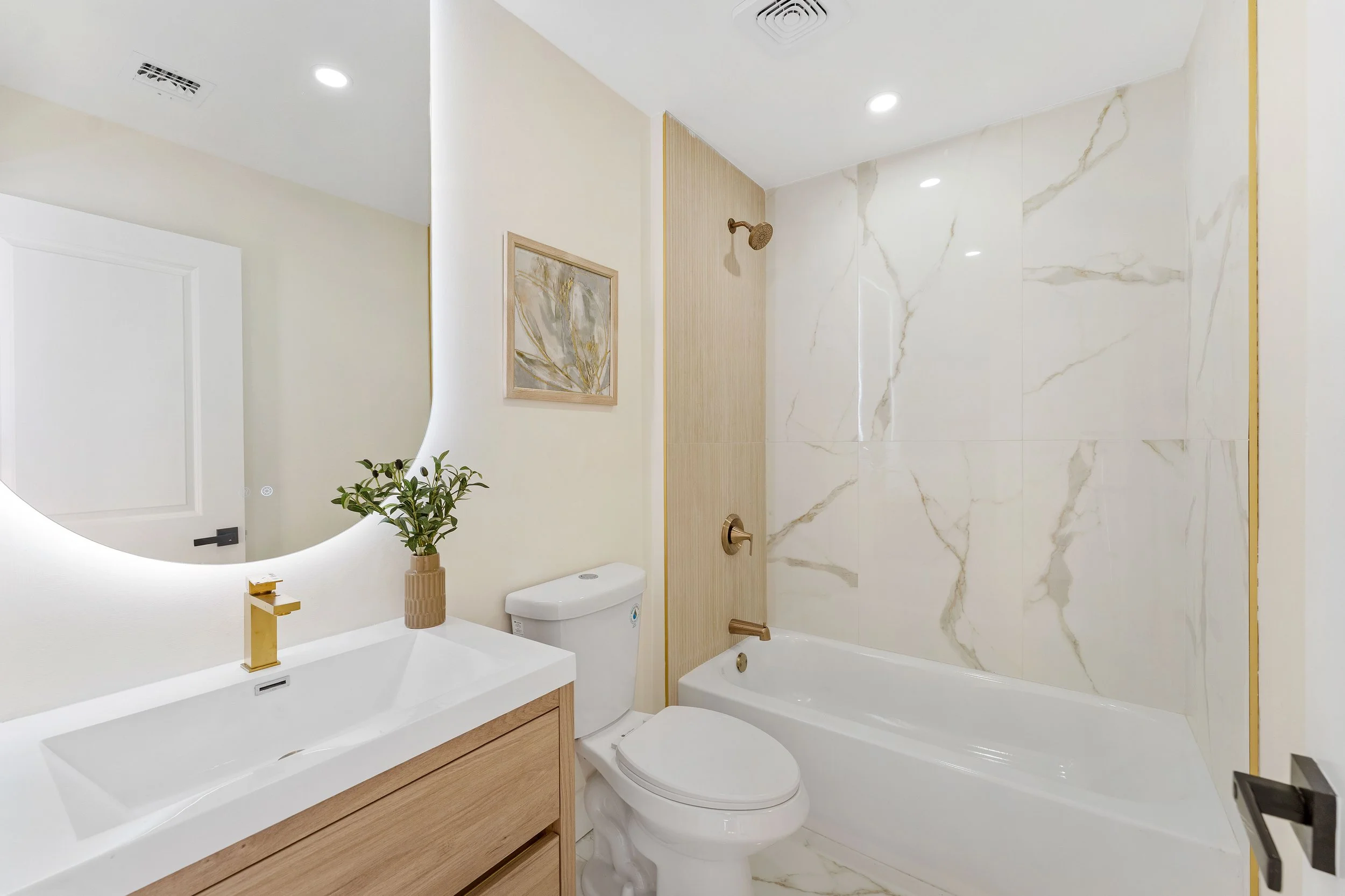 Modern bathroom with white vanity, gold faucet, potted plant, framed wall art, white toilet, marble-look shower walls, and bronze fixtures.