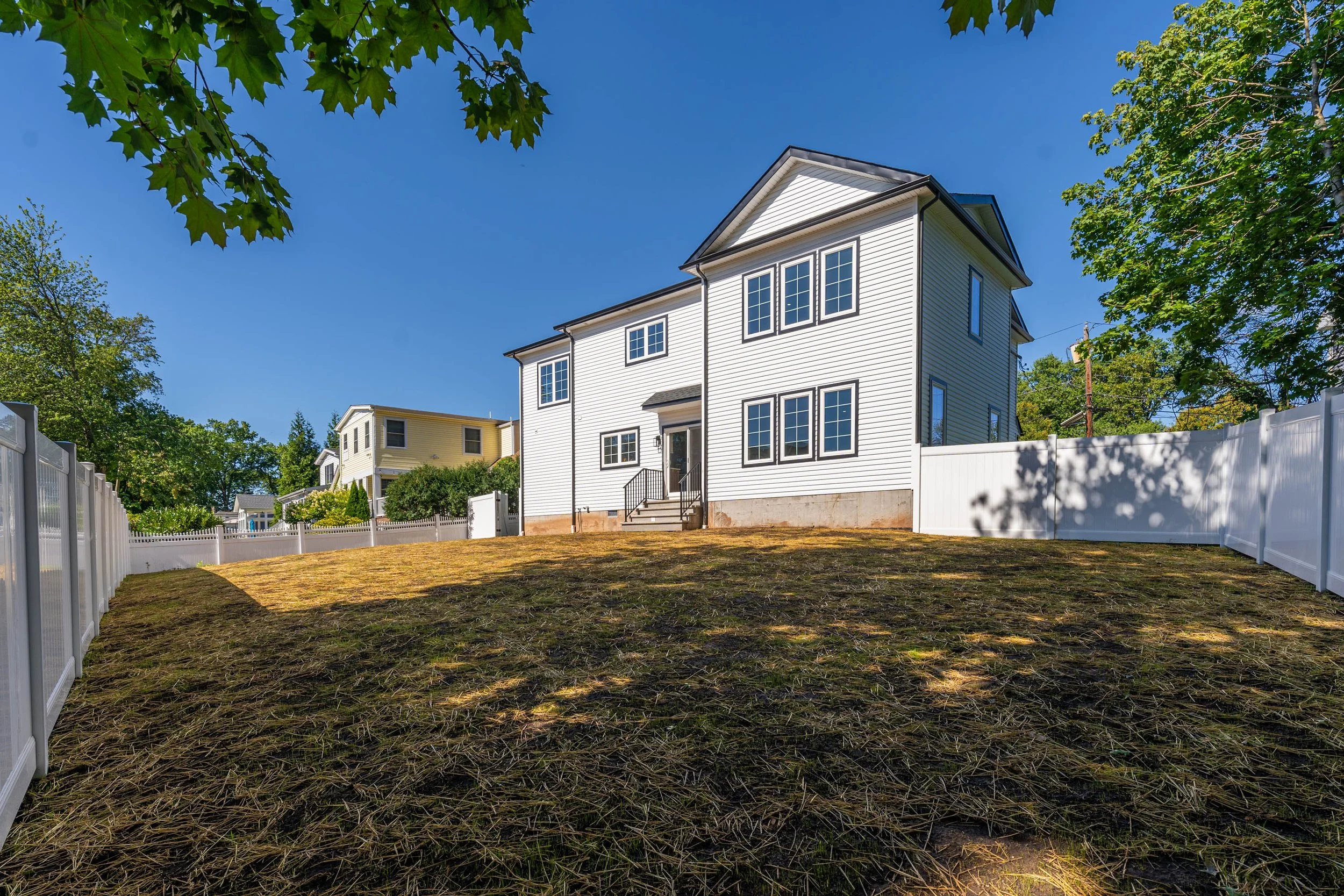 White two-story house with black window frames, steps leading to front door, fenced backyard with patchy grass, blue sky, and trees.