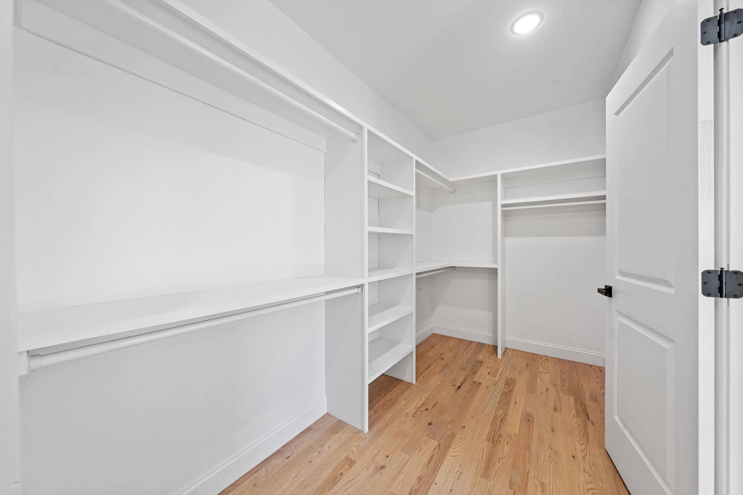 Empty walk-in closet with white shelving and hanging rods, wood flooring, and a ceiling light.