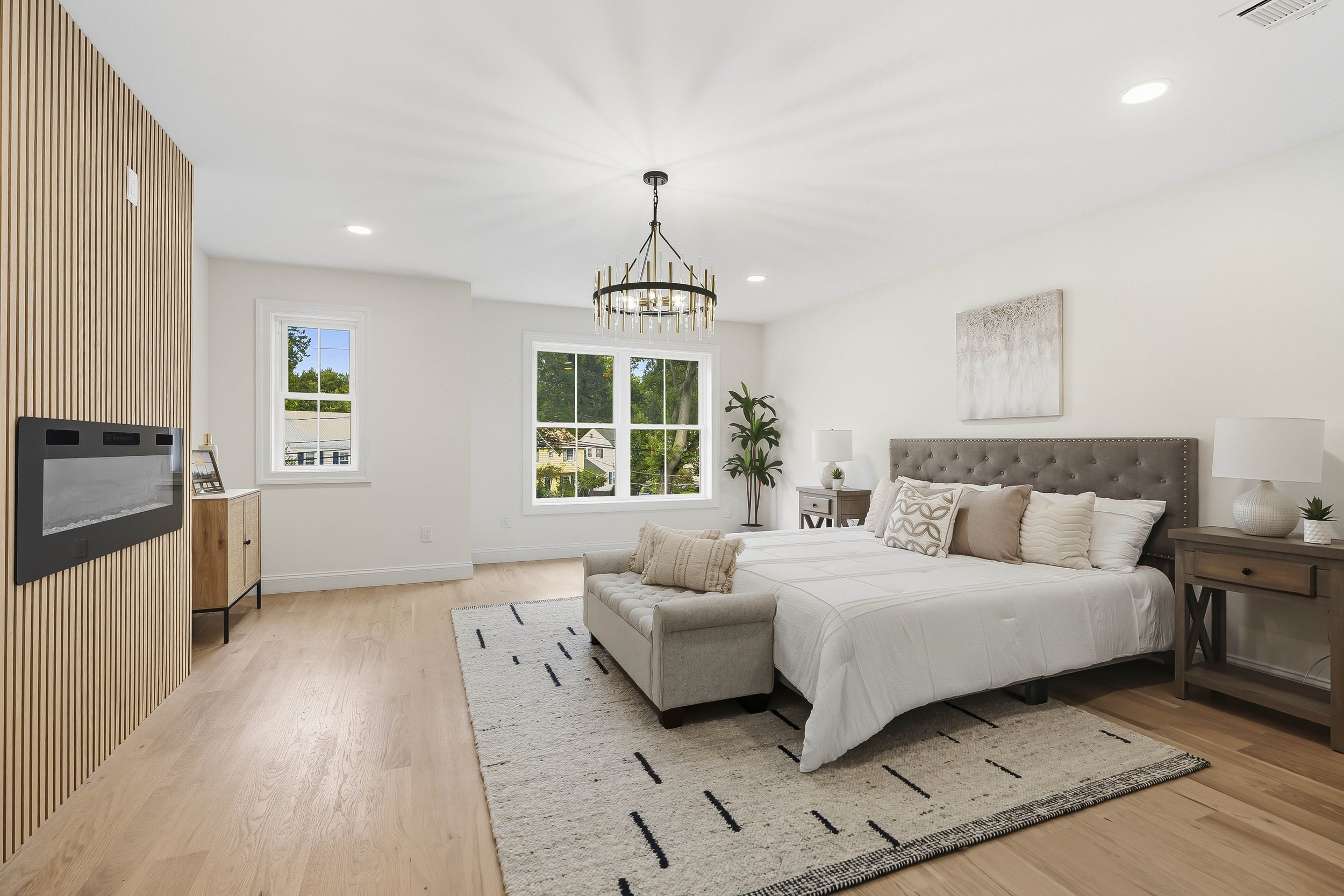 Modern bedroom with a large bed, gray tufted headboard, white bedding, two wooden nightstands with lamps, a small bench at the foot of the bed, wooden flooring, a white area rug, windows showing a view of trees and neighboring houses, and a black mod