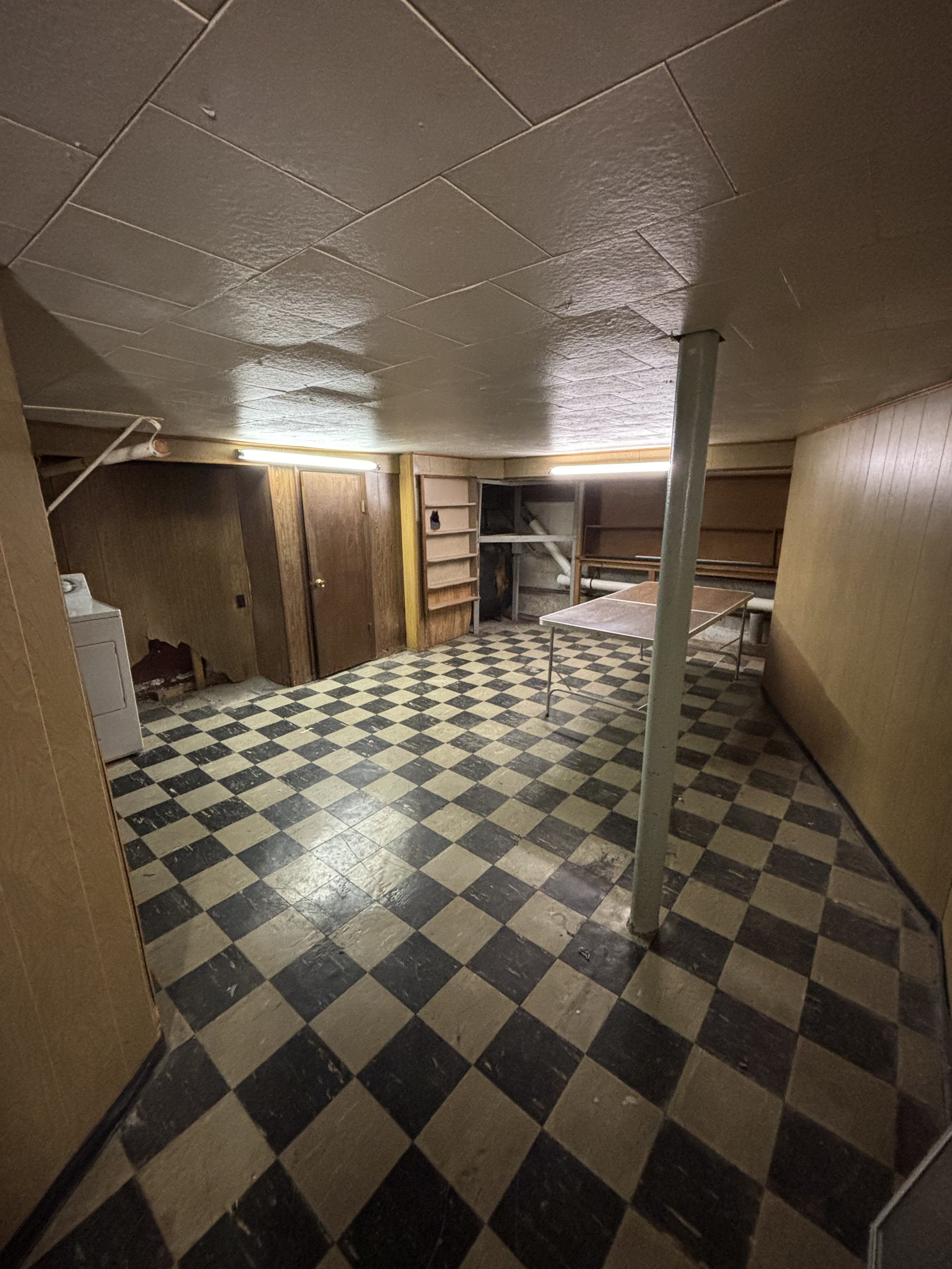 Empty basement room with checkered black and beige vinyl flooring, wooden panel walls, a metal pole in the center, and built-in wooden shelves and a door at the back. Fluorescent lighting illuminates the space.