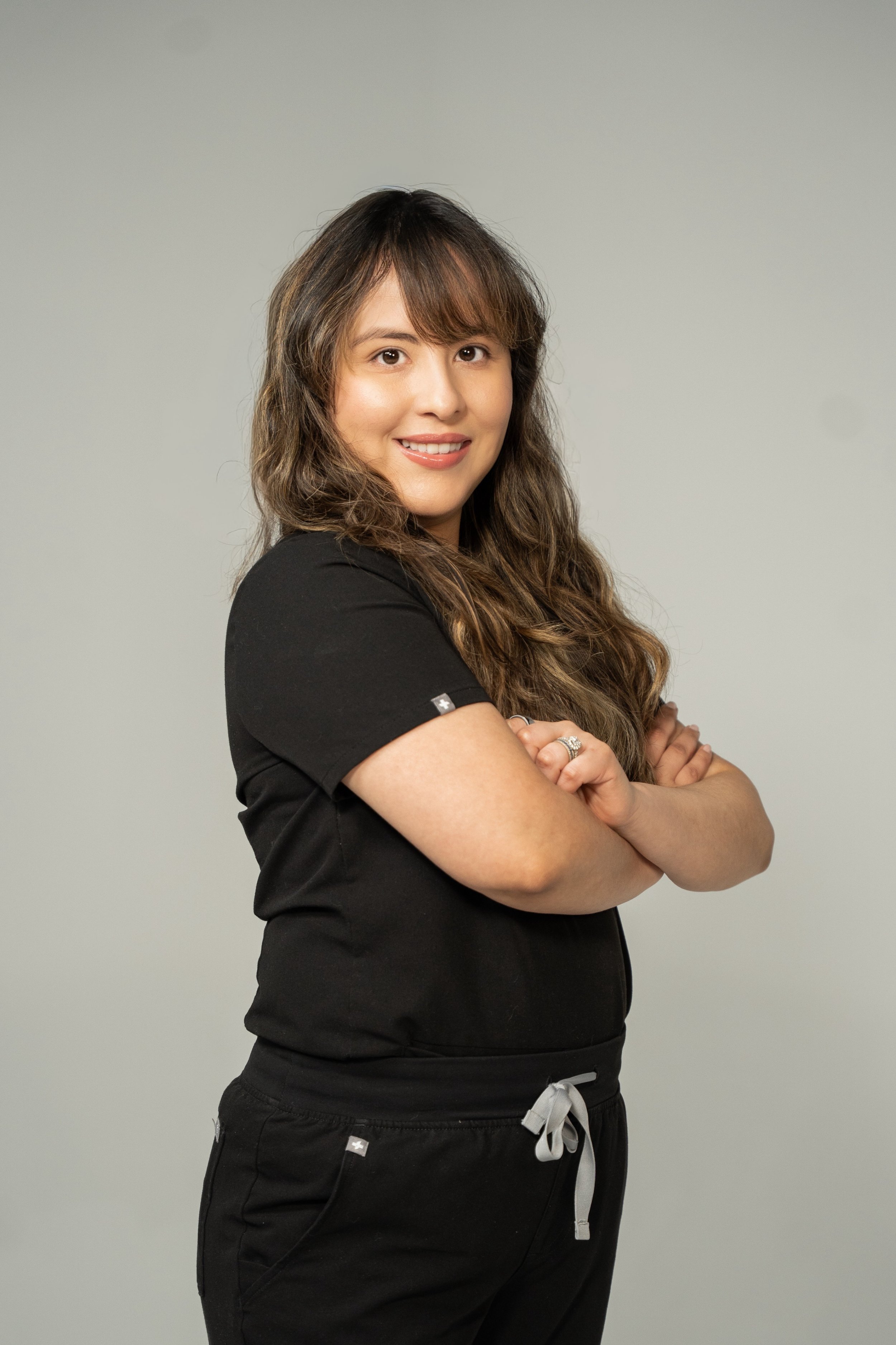 A woman with long, wavy brown hair in a black shirt and sweatpants standing with arms crossed, smiling against a plain gray background.
