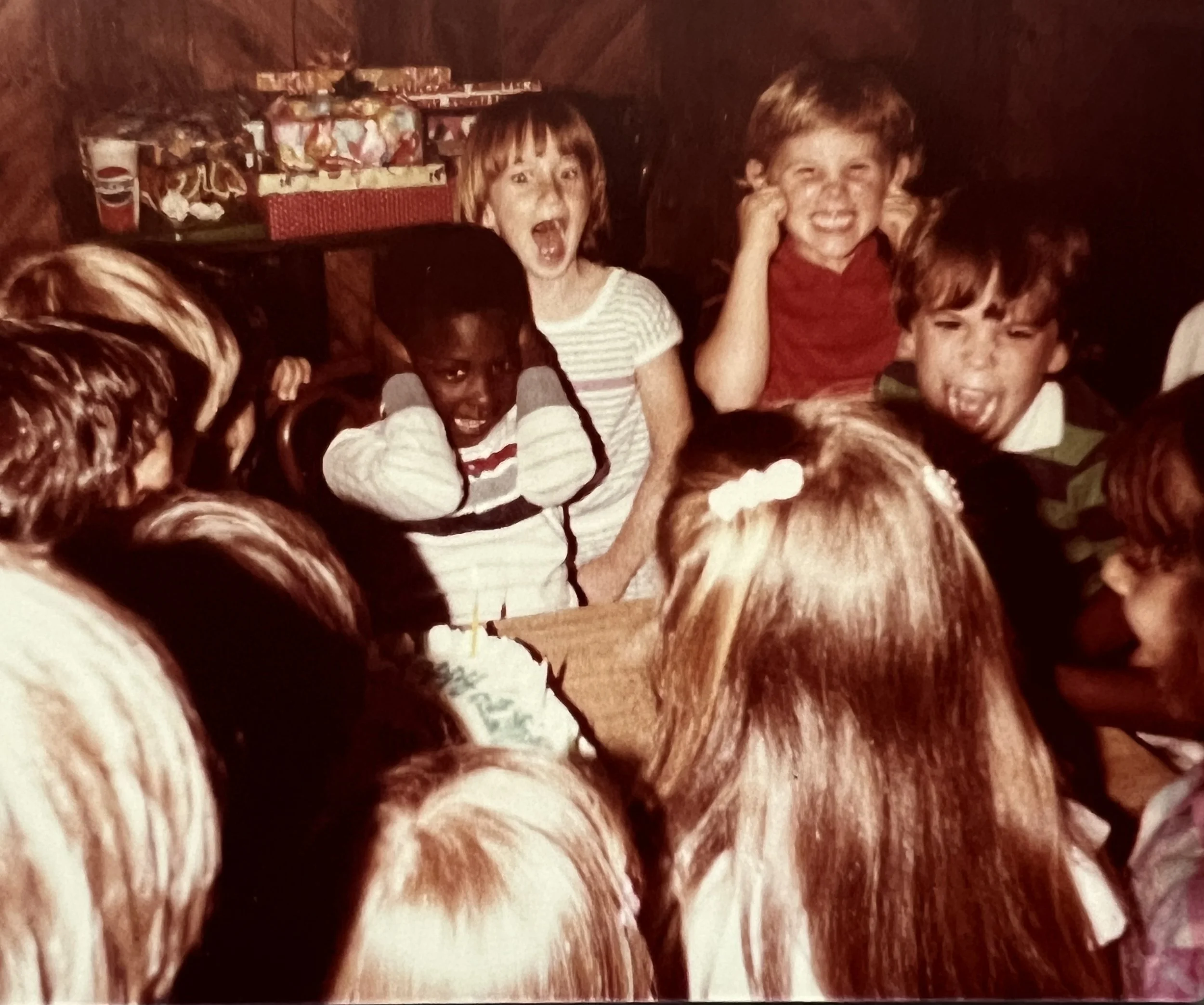 childhood picture of john during birthday party. He stands with hands covering his ears. About 9 of his friends are singing happy birthday loudly.