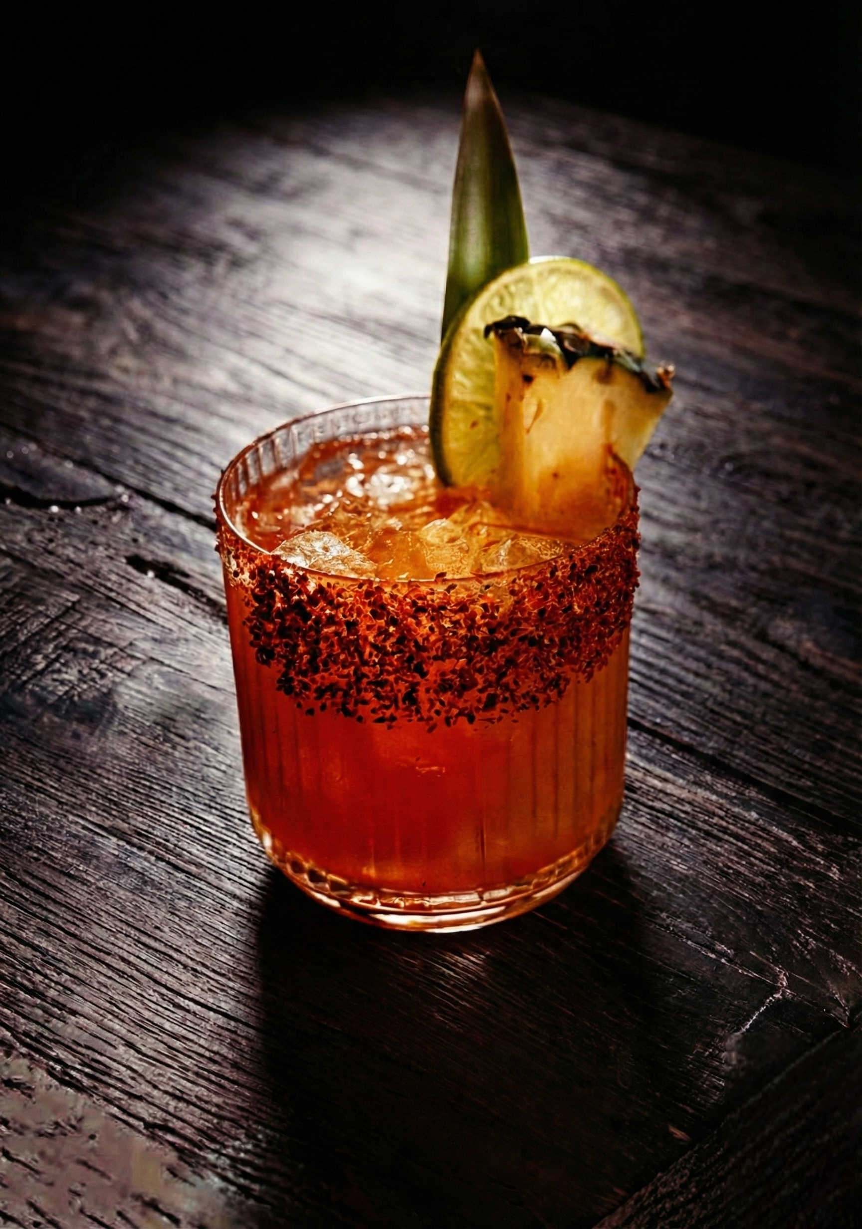 A red cocktail on the rocks with a spicy rim topped with a lemon and pineapple slice.
