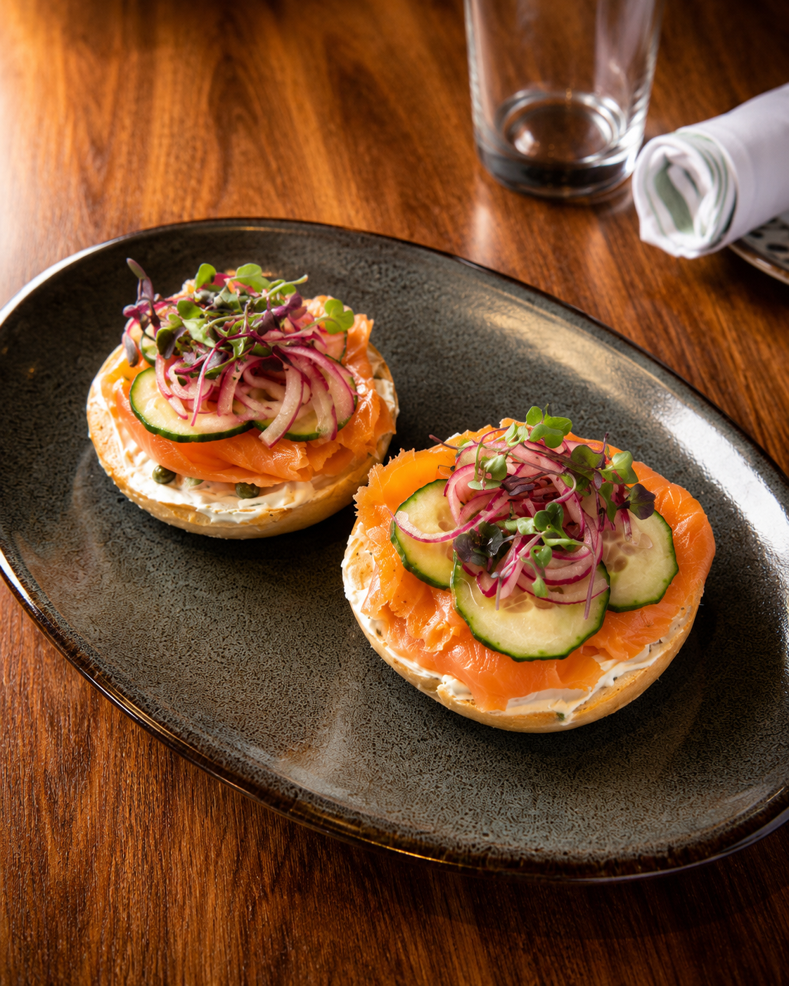 Two bagel bites topped with smoked salmon, cucumber slices, red onion, microgreens, and cream cheese on a black plate.