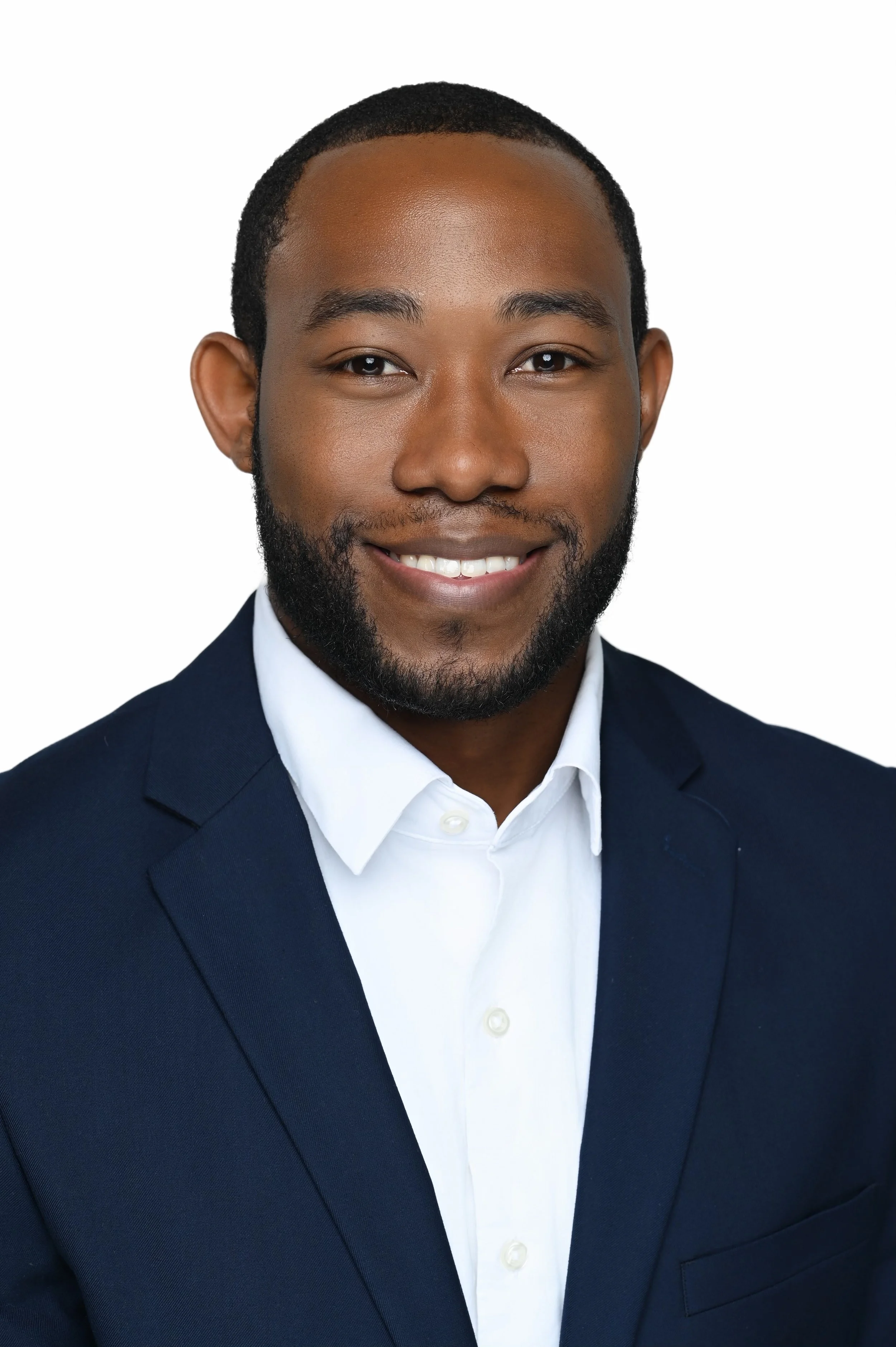 A professional headshot of an African American man with short hair and a beard, smiling, wearing a navy blue suit and white shirt, against a white background.
