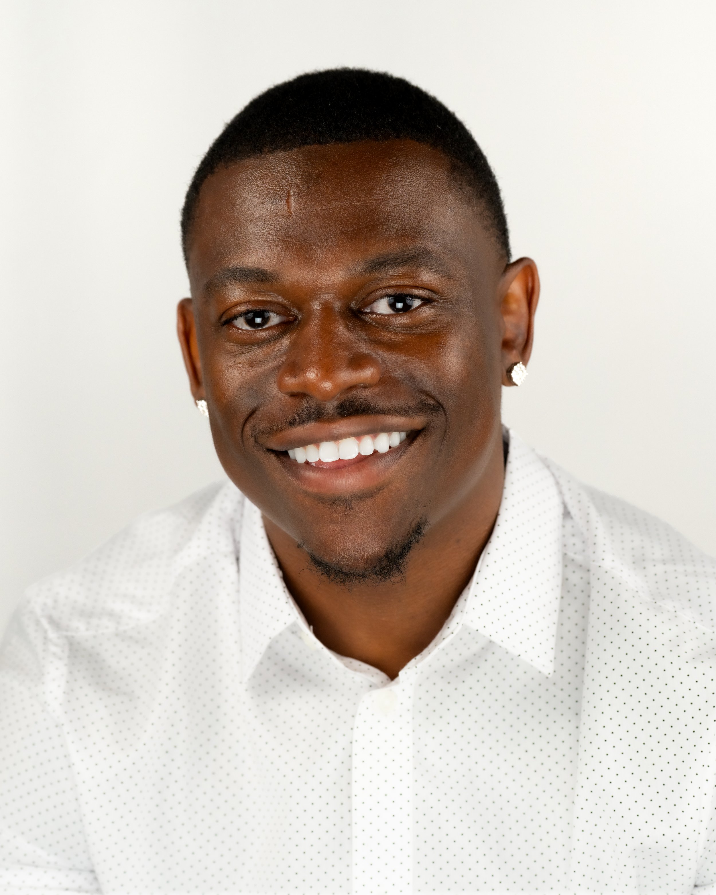 Headshot of a smiling Black man with short hair, wearing earrings and a white shirt with small polka dots, against a plain white background.