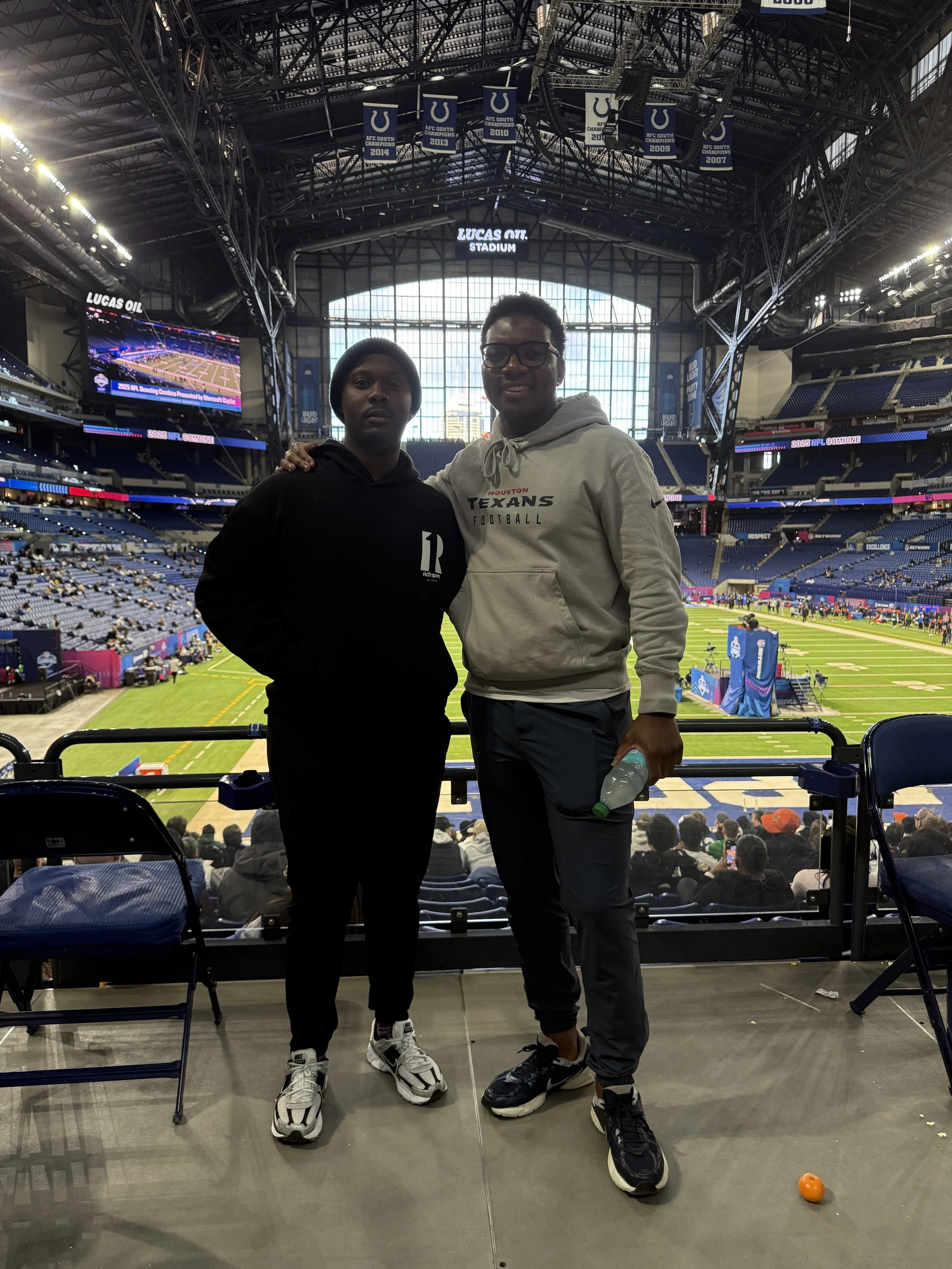 Two young men standing inside a large football stadium, with the field visible behind them. The stadium has banners hanging from the ceiling and many spectators in the stands. They are posing for the photo, dressed in casual sportswear.