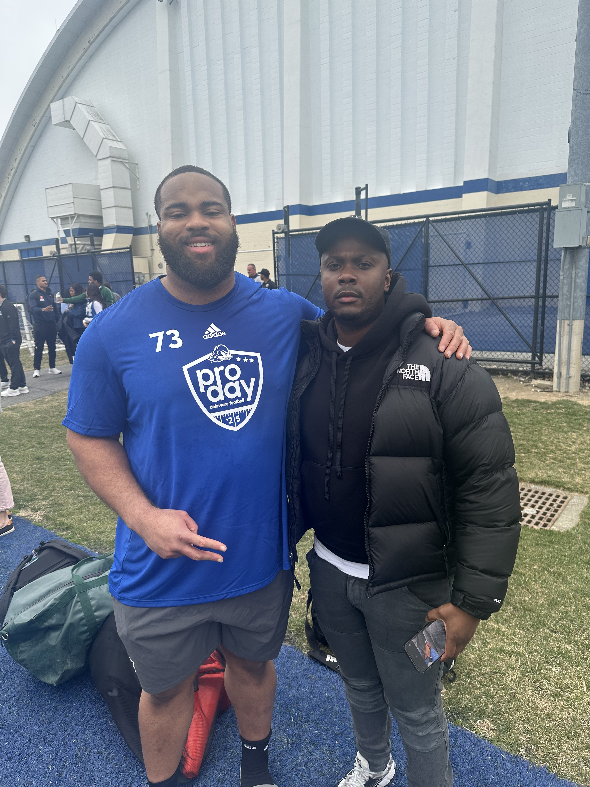Two men posing outdoors, one wearing a blue pro day shirt and the other wearing a black North Face puffer jacket, with a group of people in the background at a sports facility.
