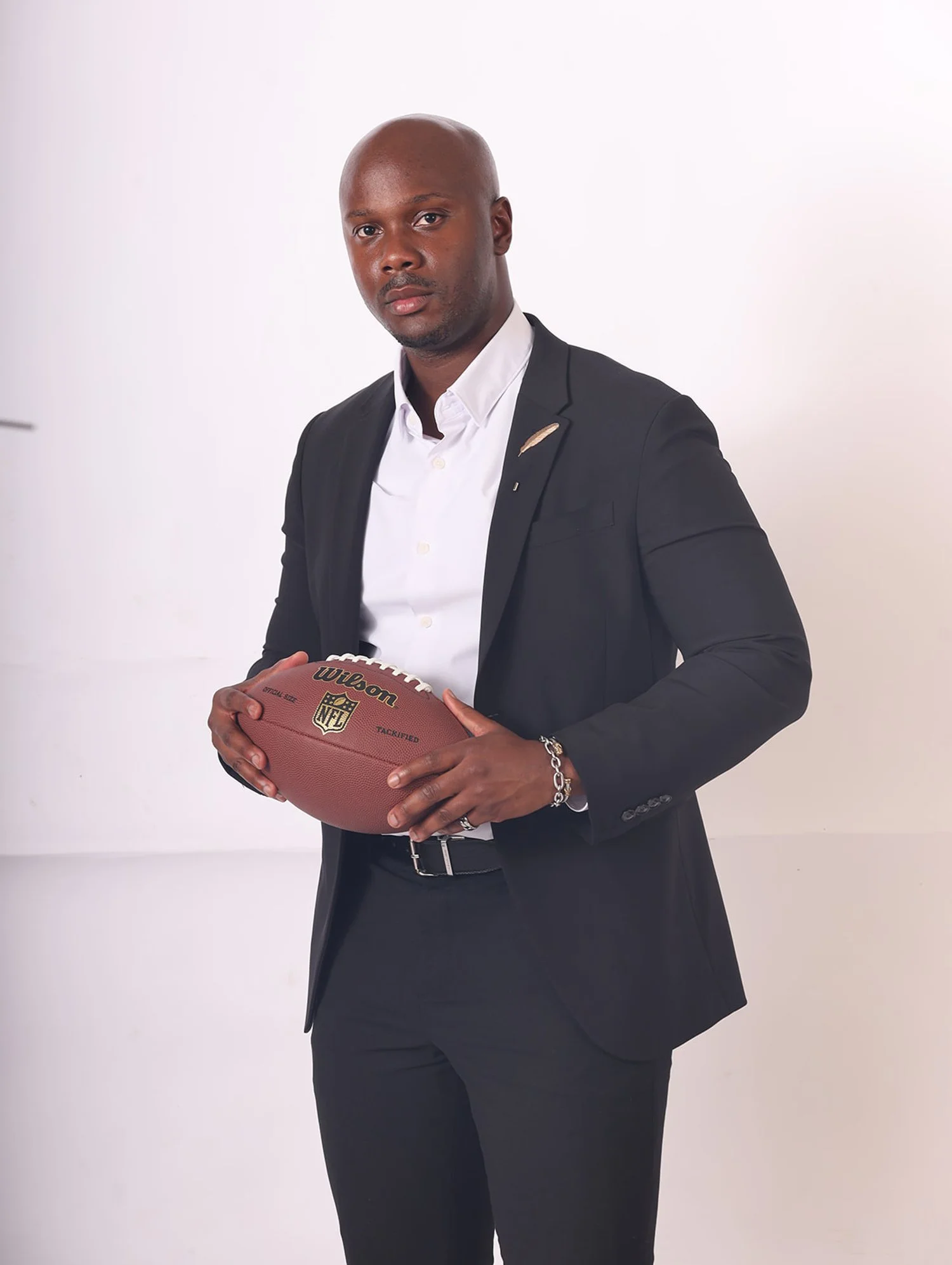 A man in a black suit and white shirt holding a football against a plain white background.