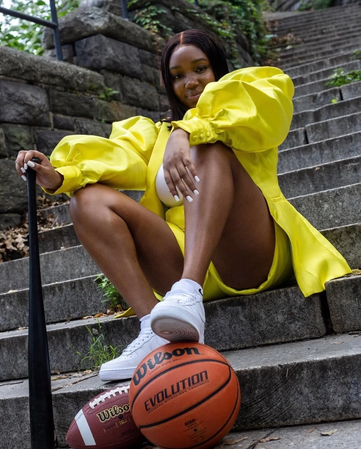 A woman wearing a bright yellow jacket and shorts sitting on outdoor stone stairs. She has white sneakers, and two basketballs are in front of her, one Wilson and one Evolution. She has dark hair and is looking directly at the camera, with one knee raised and her hand resting on her leg.