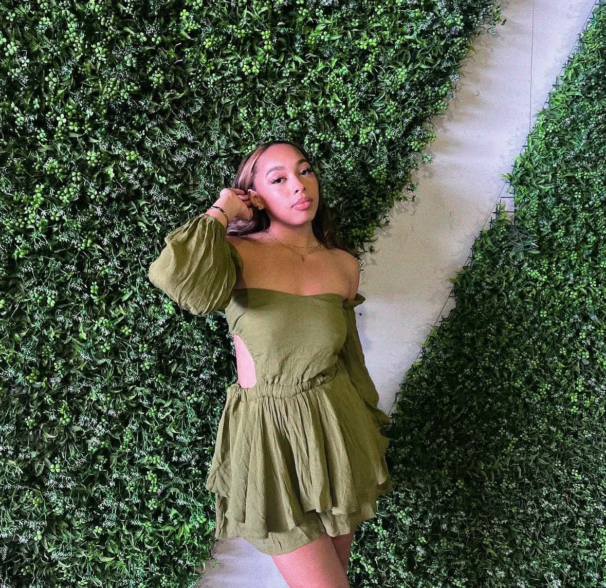 A young woman in an olive-green off-the-shoulder dress with cutouts on the sides poses against a backdrop of lush green foliage.