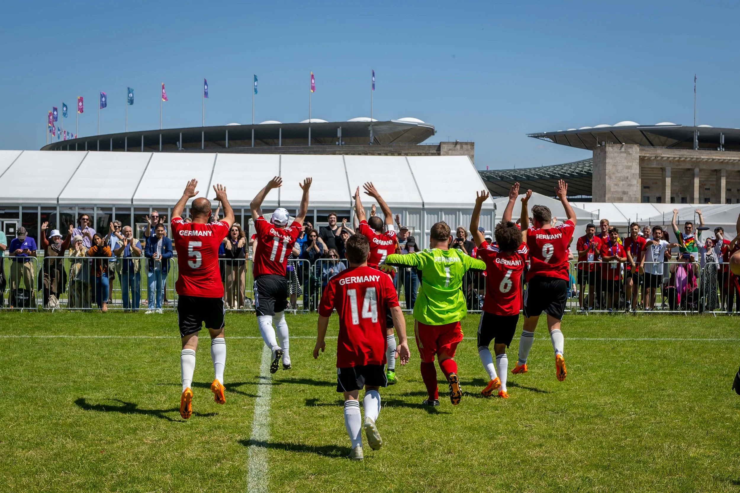 Das deutsche Fußballteam feiert sich selbst auf dem Maifeld. Im Hintergrund stehen jubelnde Fans, über ihnen strahlt der blaue Sommerhimmel. Temporäre Zeltbauten stehen vor dem Berliner Olympiastadion.
