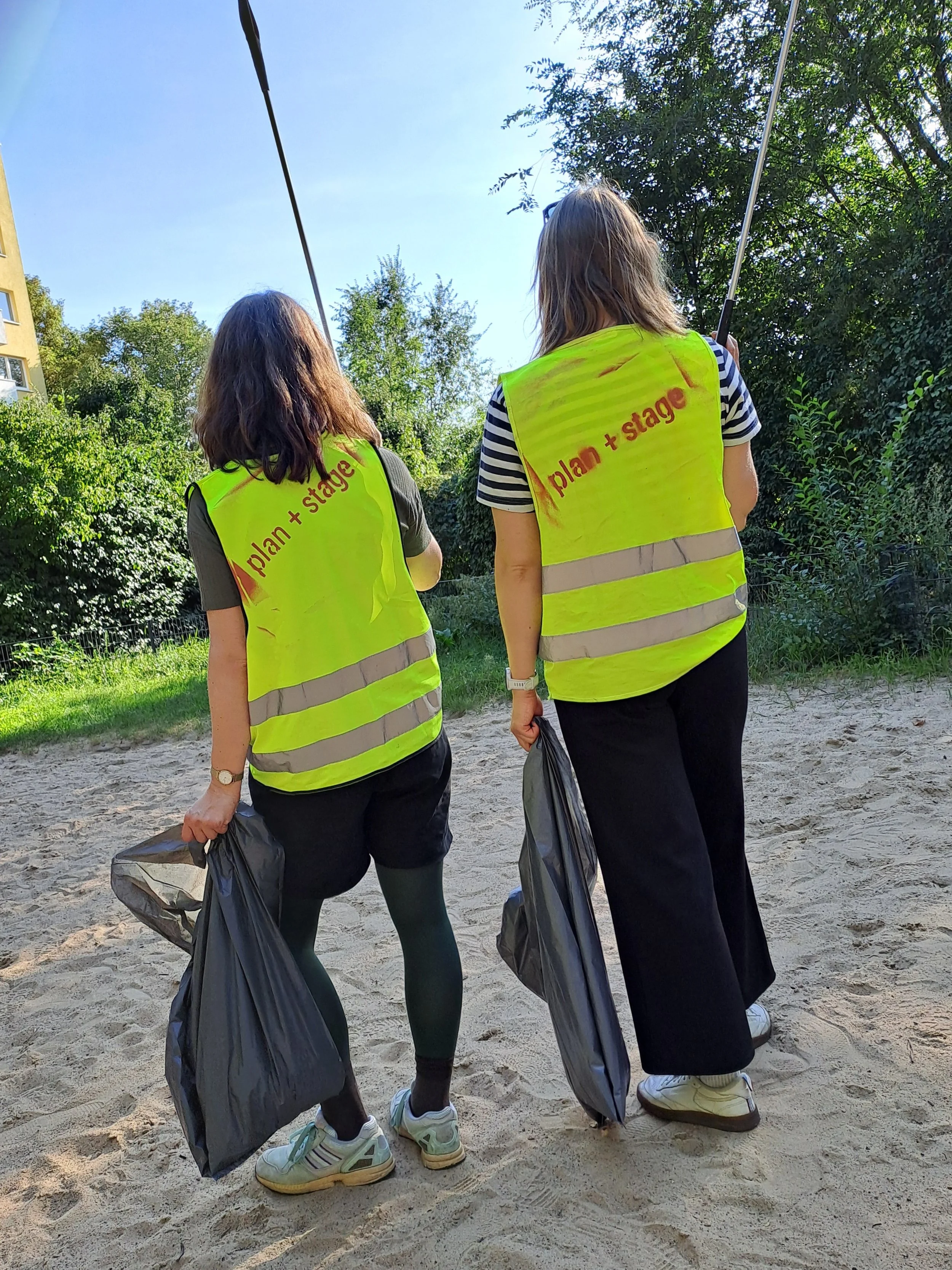 Zwei Mitarbeiter*innen von plan + stage tragen gelbe Warnwesten und sammeln Müll.
