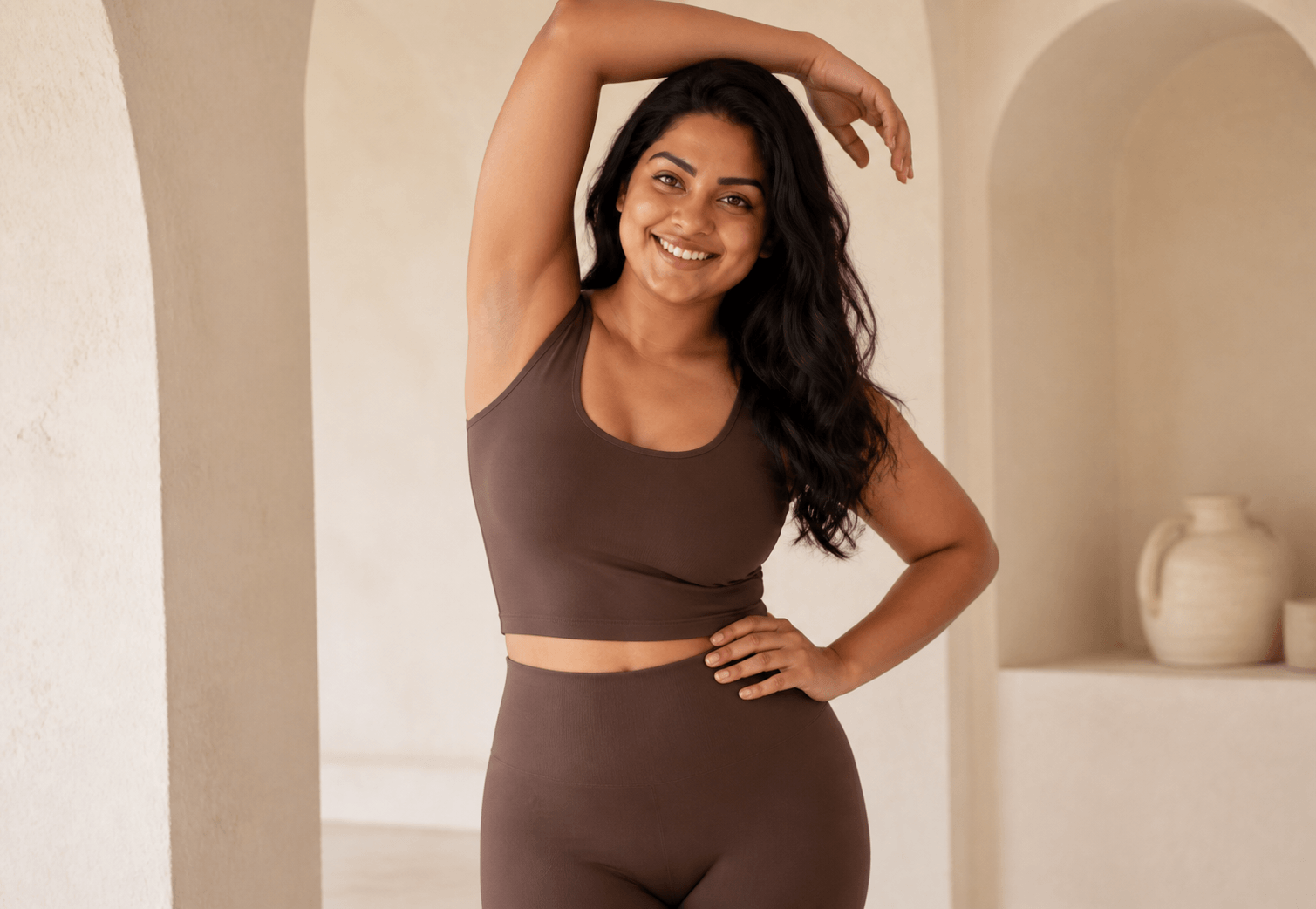 A woman with long dark hair smiling and posing in a brown workout outfit in a neutral-colored room.