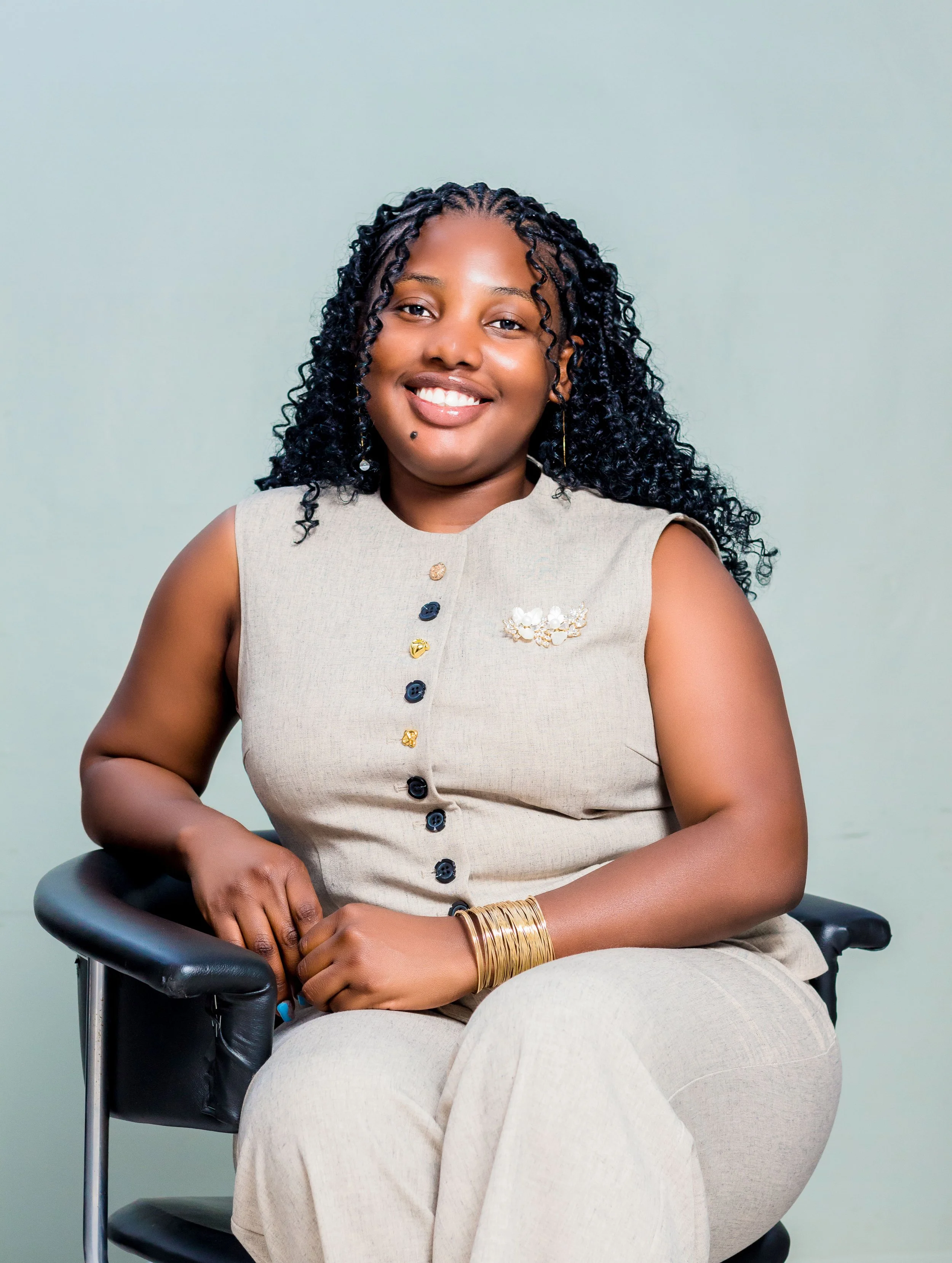 A woman with dark, curly hair wearing a beige sleeveless top with decorative buttons, sitting on a black chair with a light green background.