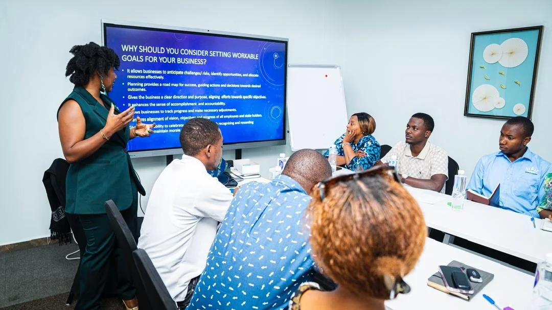 A woman presents to a group of diverse professionals in a conference room, standing next to a large screen displaying a presentation titled 'Why should you consider setting workable goals for your business?' The attendees are seated around a table, listening attentively.