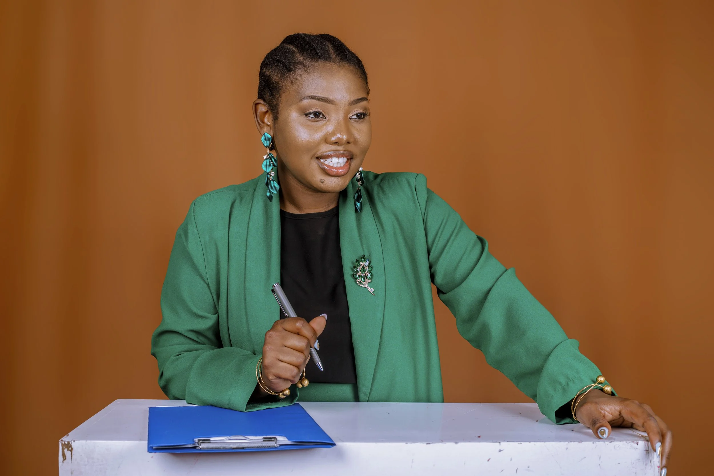 A woman sitting at a table holding a pen, wearing a green blazer with blue earrings and a brooch, with a brown background.