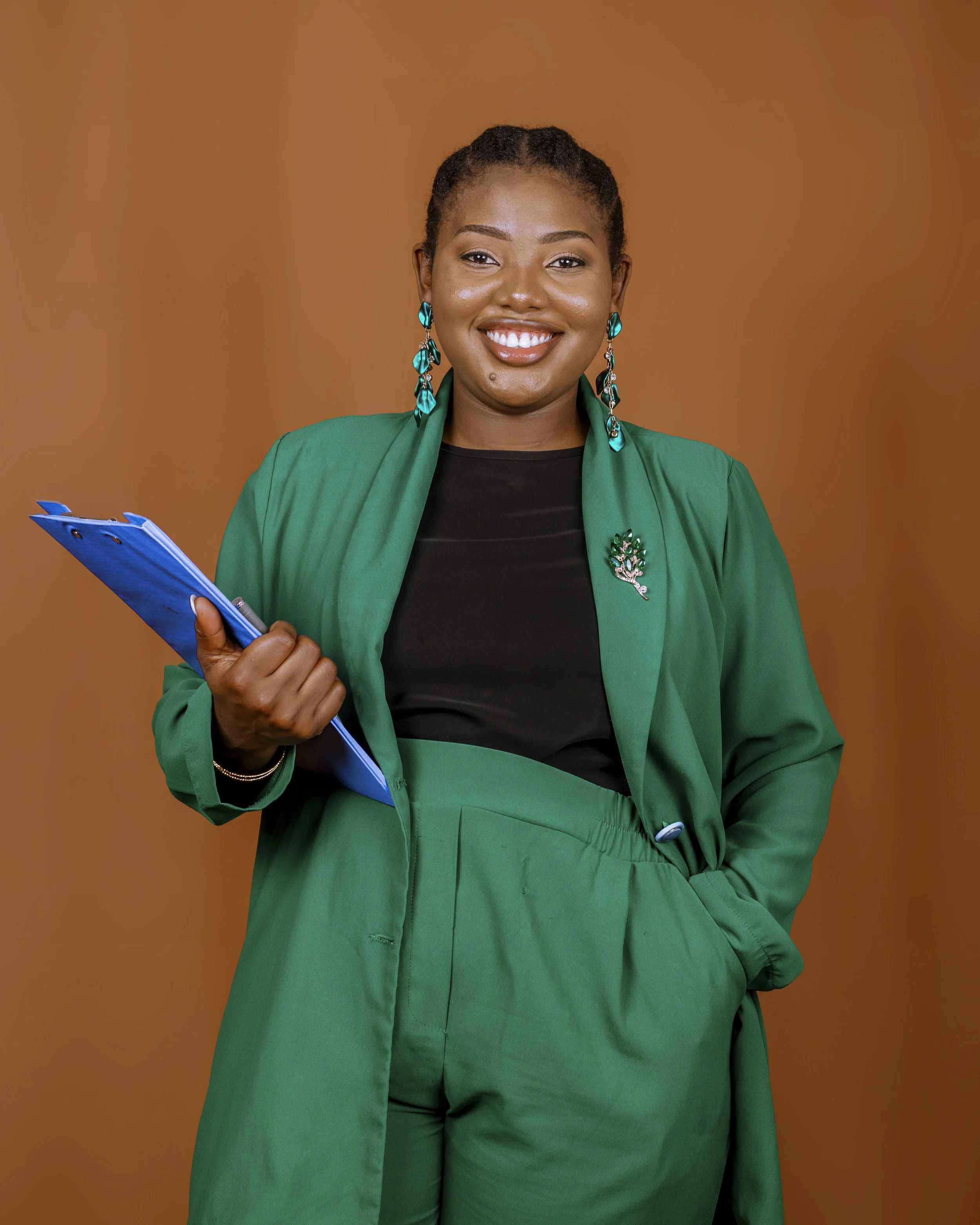 Smiling woman wearing green blazer and matching pants, holding a blue clipboard, standing against an orange background.