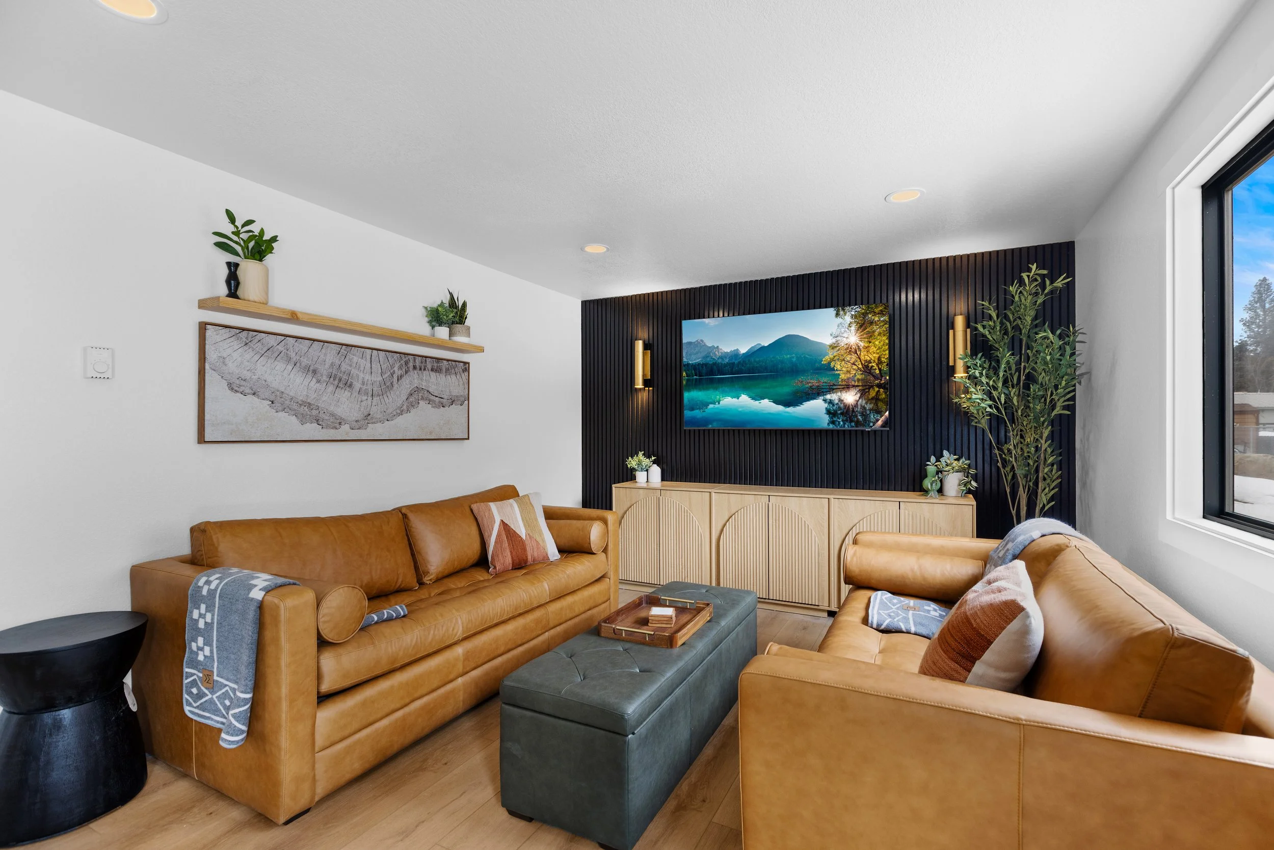 Living room with two leather sofas, a black ottoman, wall-mounted flat screen TV on a black panel, wooden sideboard, framed artwork, floating shelves with plants, large window showing outdoor view, and indoor plants.