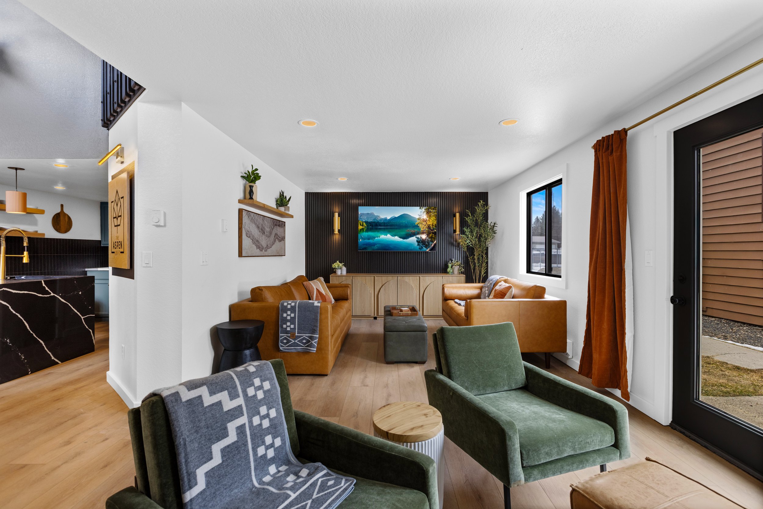 Living room with two tan sofas, green armchairs, and a wall-mounted TV displaying a lake scene. The room has white walls, light wood flooring, and large windows with brown curtains, overlooking an outdoor space.