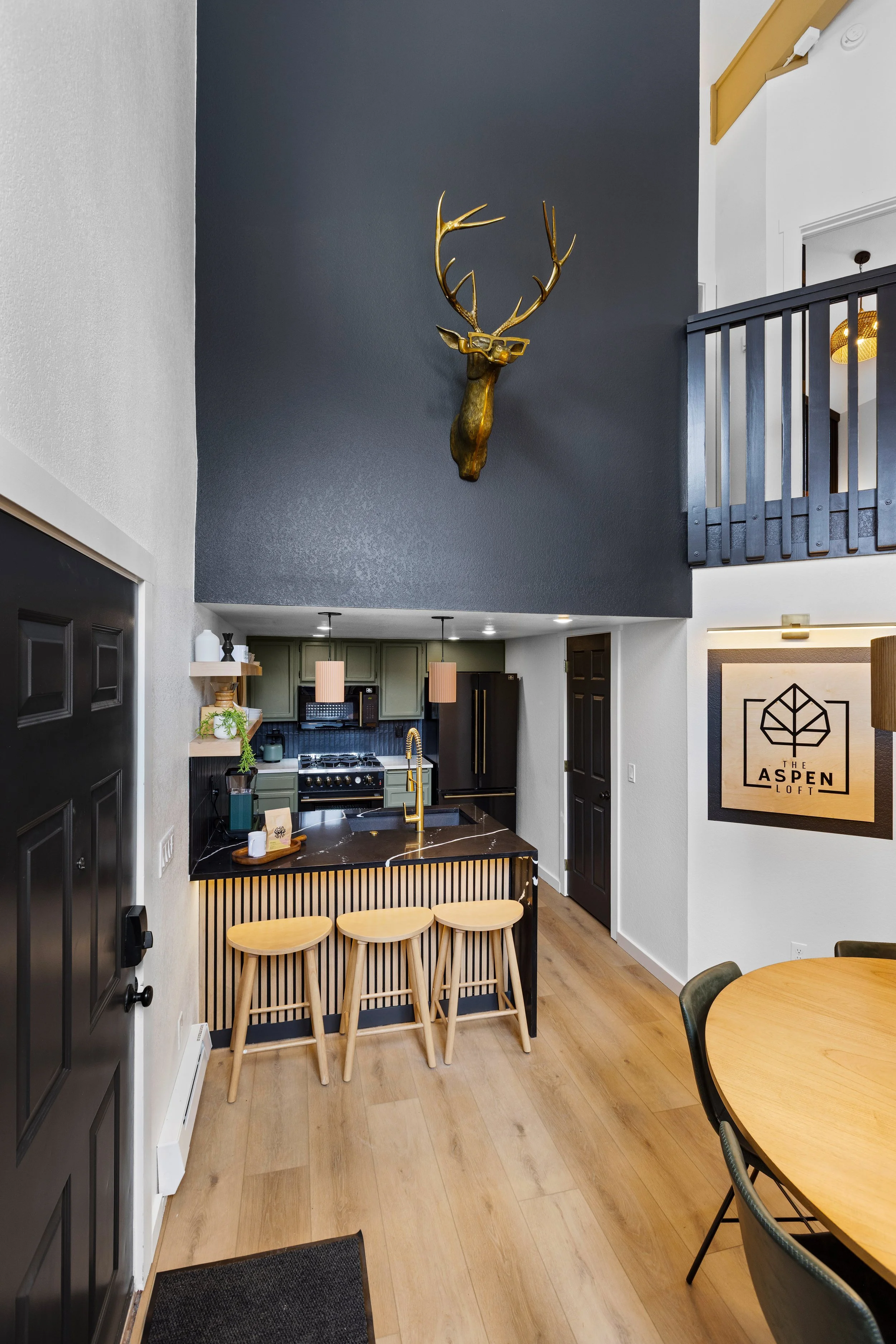 Interior view of a modern apartment kitchen and dining area with a high dark-colored accent wall, a mounted deer head with glasses and antlers, a black kitchen counter with three wooden stools, green cabinets, a black refrigerator, and a round wooden