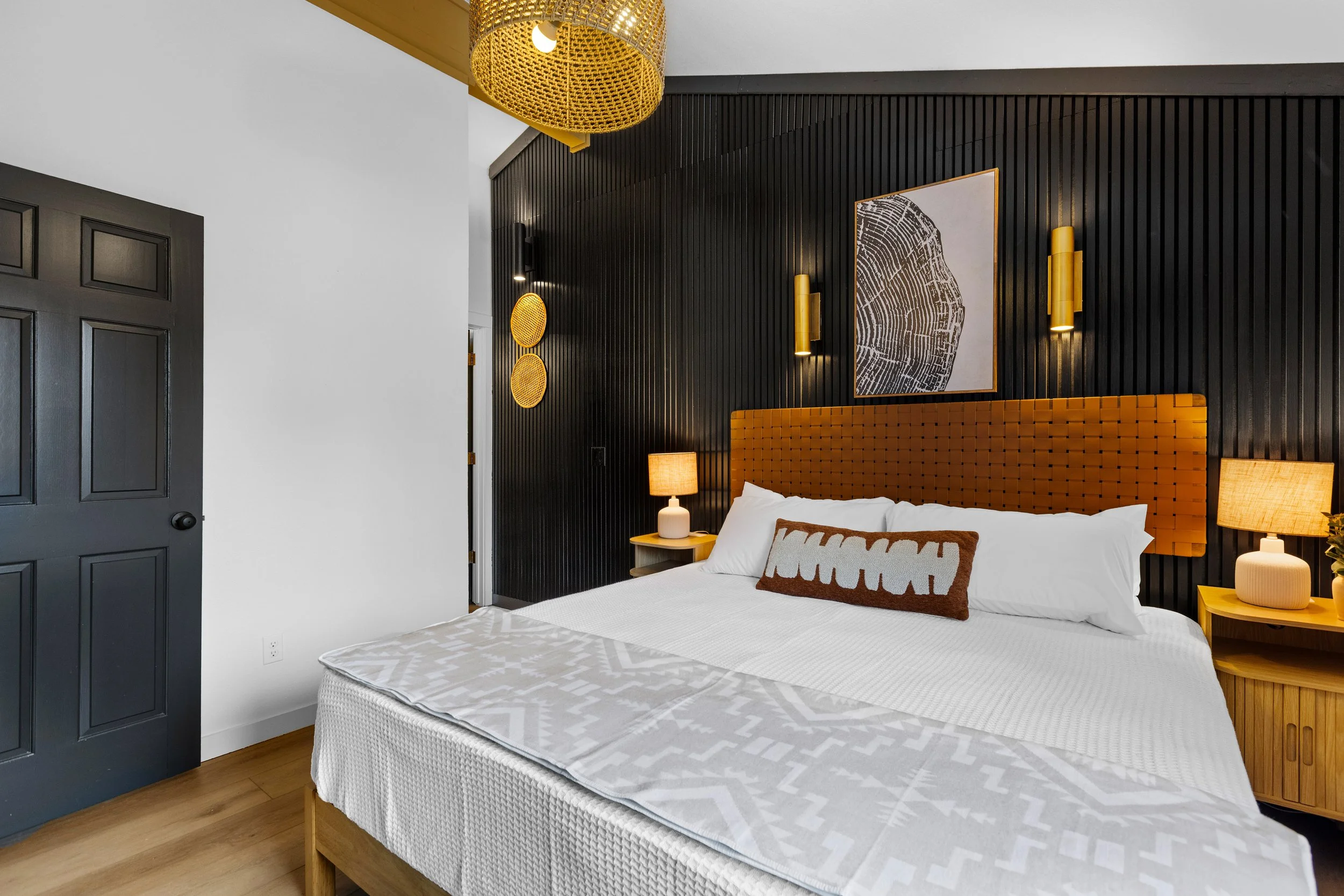 Modern bedroom featuring a bed with white bedding and a decorative pillow, black textured accent wall with wall-mounted lights, framed artwork, bedside tables with lamps, and wood flooring.