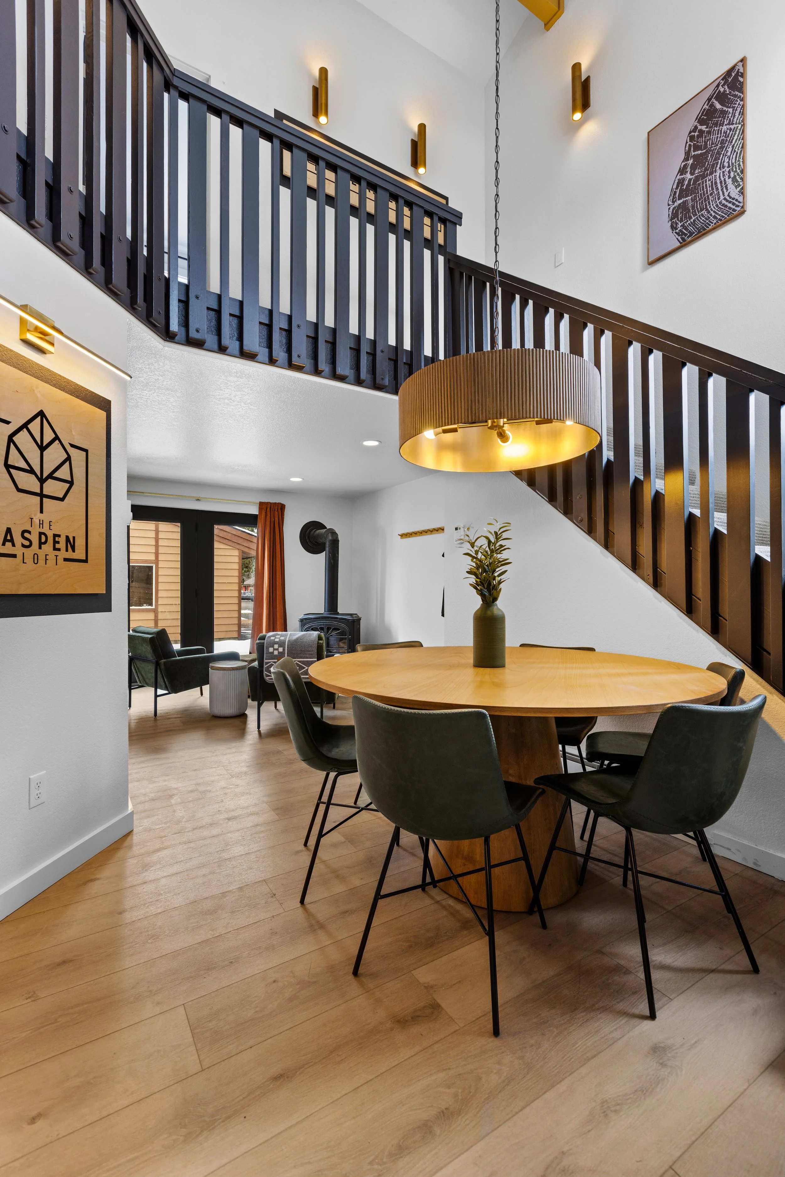 Interior of a modern living space with a high ceiling, featuring a round wooden dining table with six black chairs, a large hanging light fixture, and a staircase with dark wood railings. There are windows with brown curtains, artwork on the walls, a